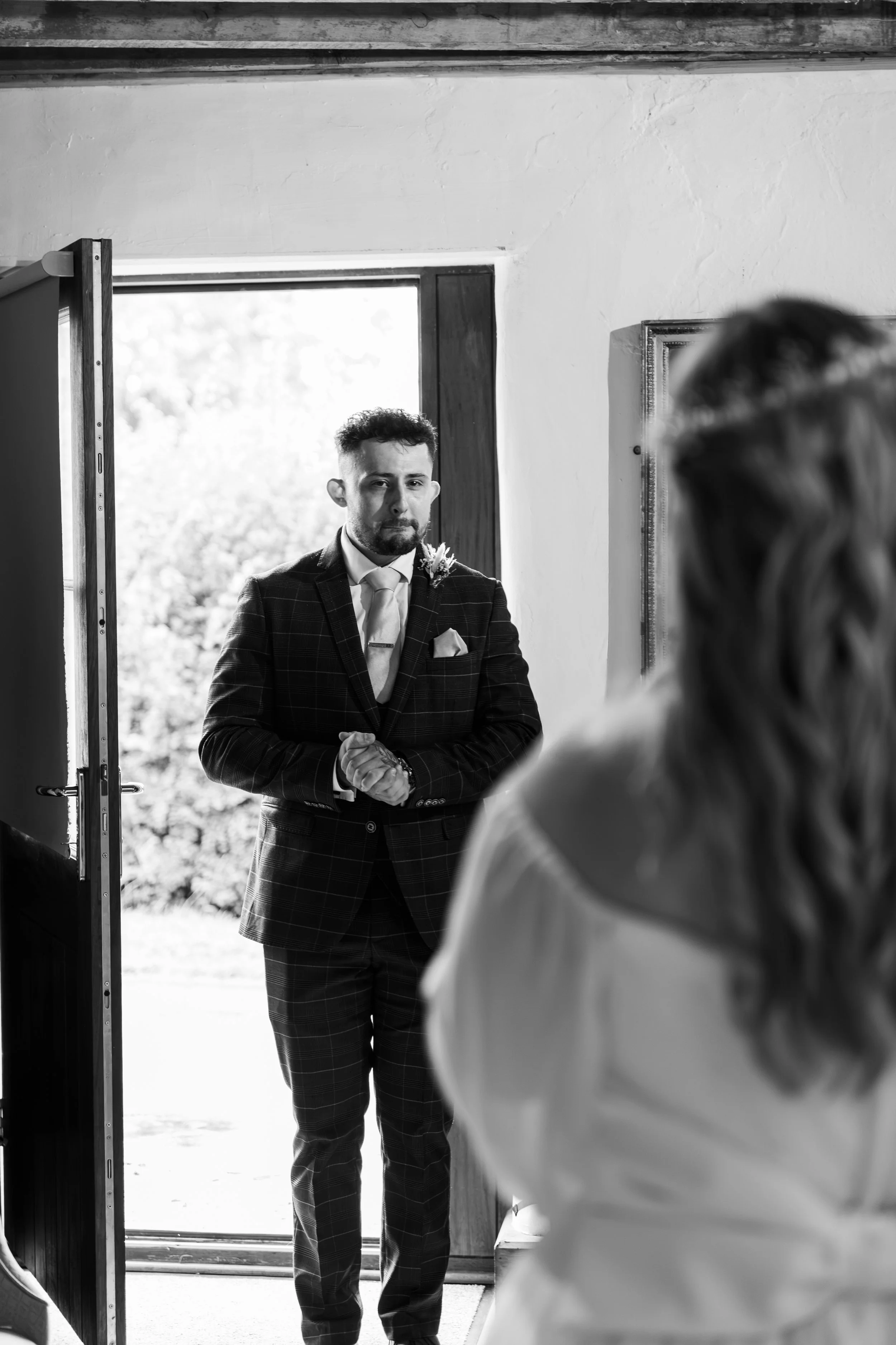 A man wearing a suit and tie standing at an open door, greeting or waiting for a woman in a white coat who is partially visible in the foreground, inside a bright room.