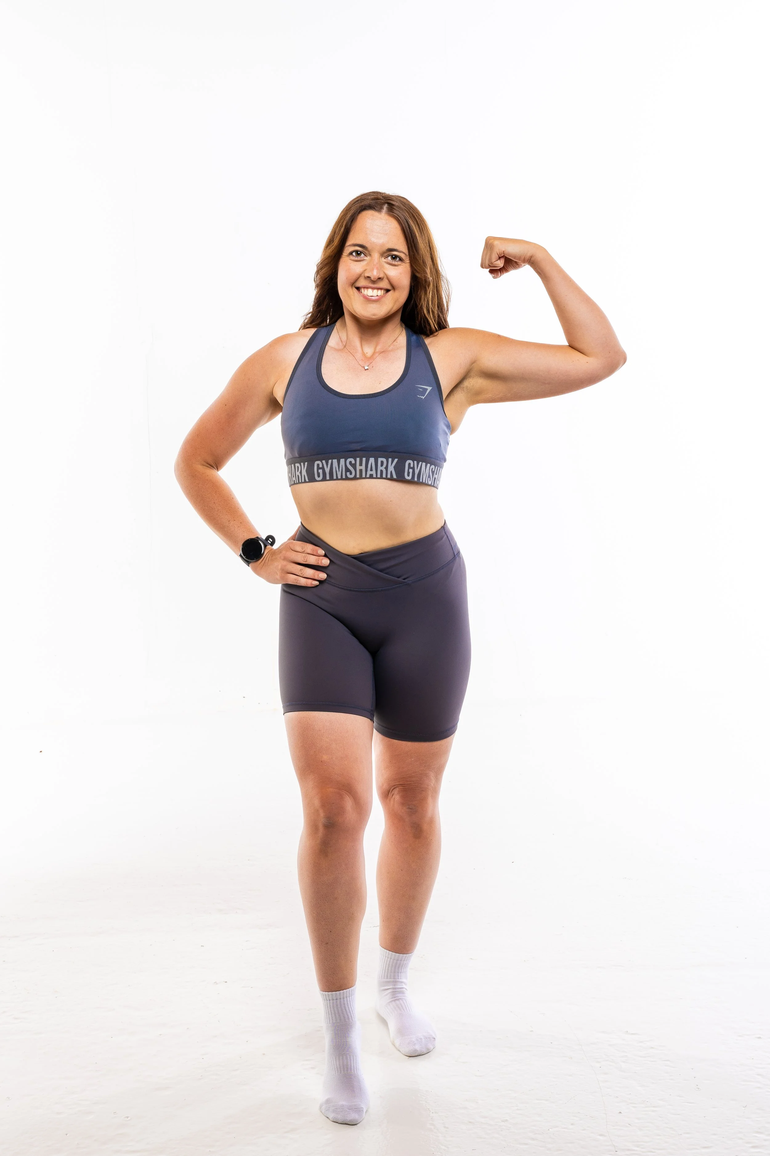 A woman with brown hair smiling and flexing her right arm, wearing workout clothes, standing in front of a plain white background.