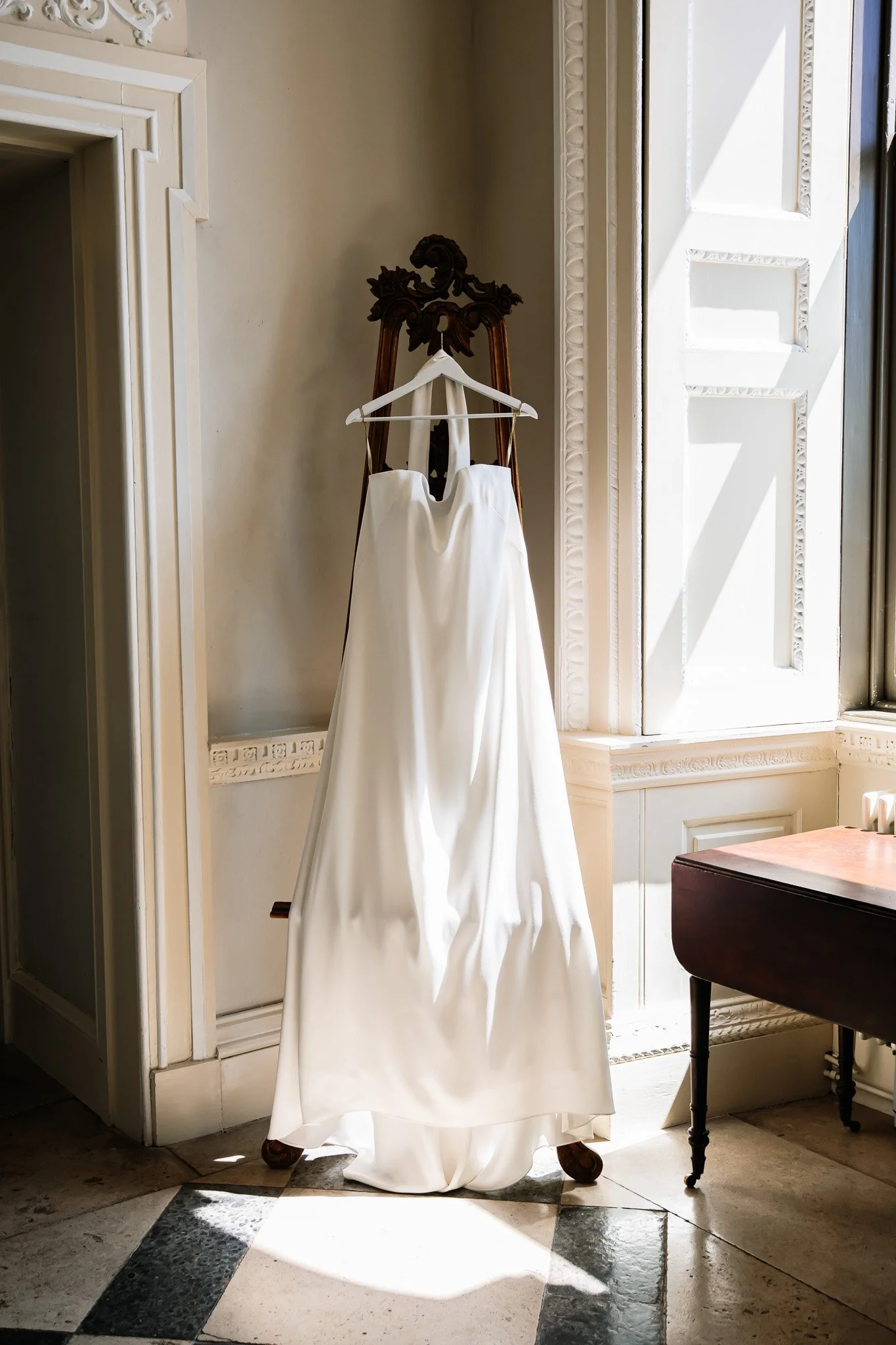 White wedding dress hanging on a hanger, draped over a wooden chair in a well-lit room with ornate white wall moldings and a window with sunlight pouring in.