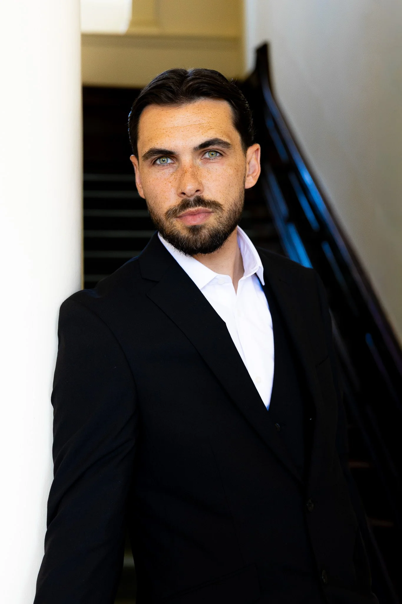 A man in a black suit and white shirt leaning against a white wall, with stairs or a staircase in the background.