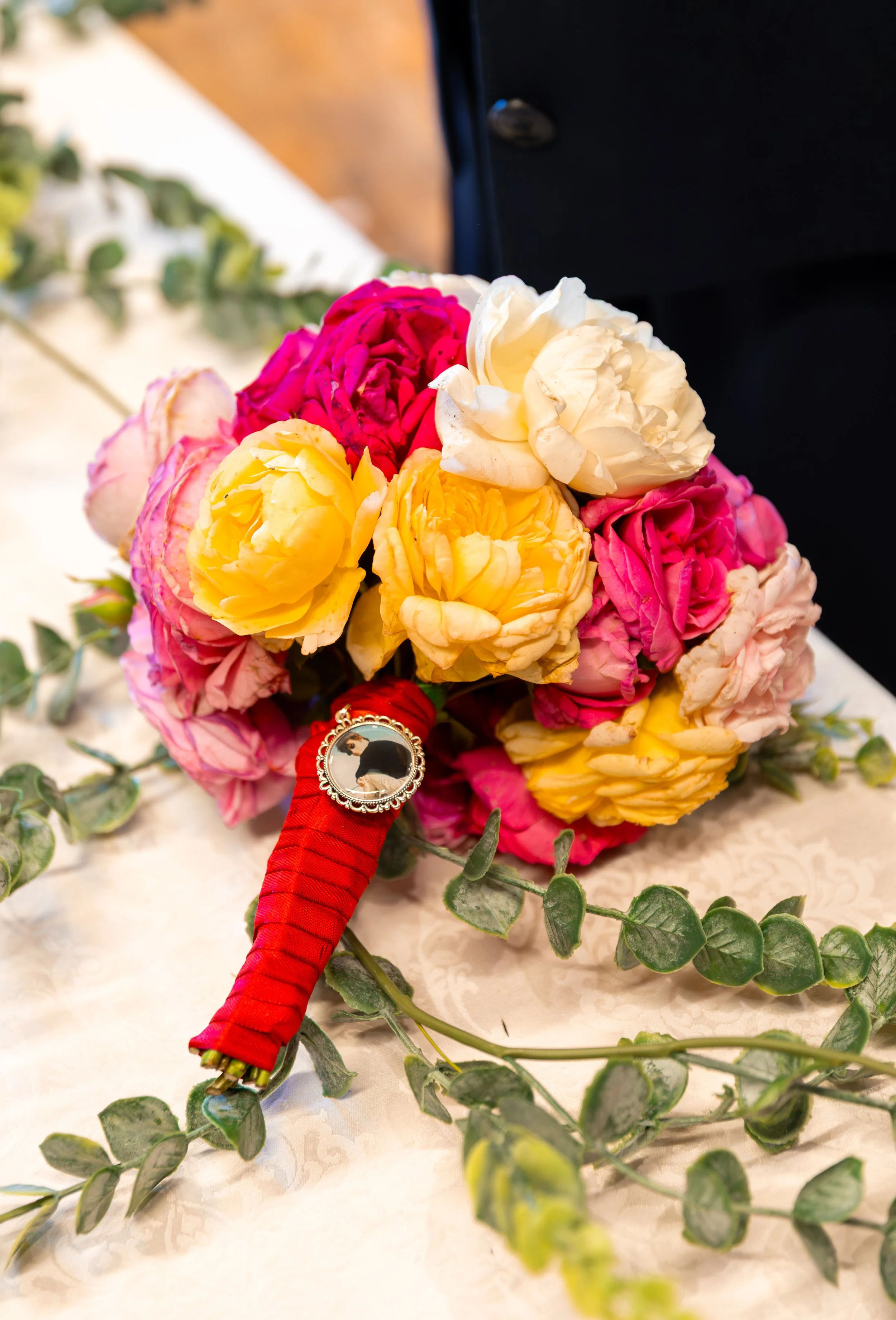 A bouquet of colorful roses, including white, yellow, pink, and red, with green leaves and eucalyptus, placed on a cream-colored lace tablecloth.