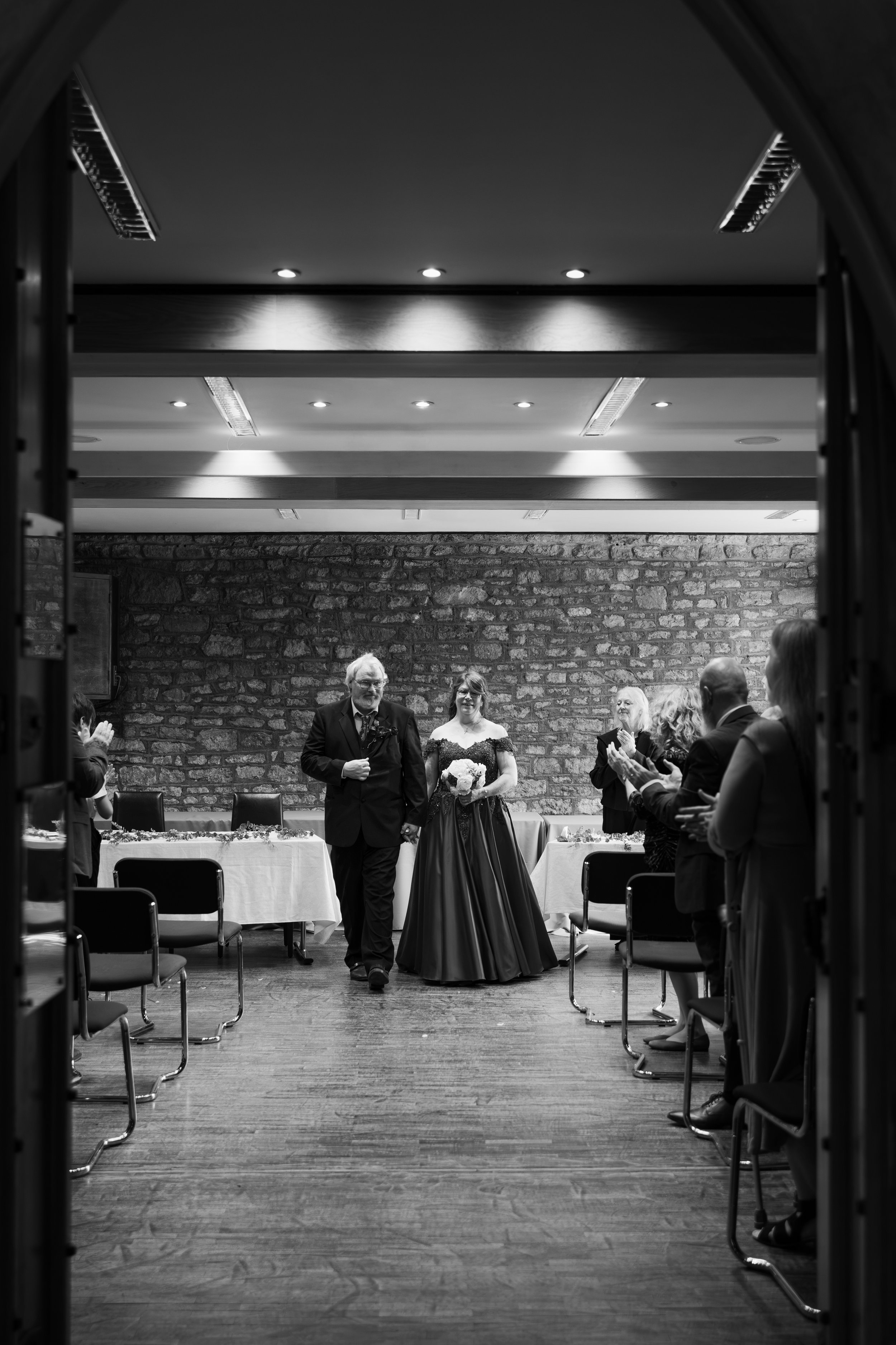 A couple dressed in formal attire walking hand in hand through a banquet hall decorated for a wedding or celebration. Guests are standing and clapping along the sides of the room as the couple makes their entrance.