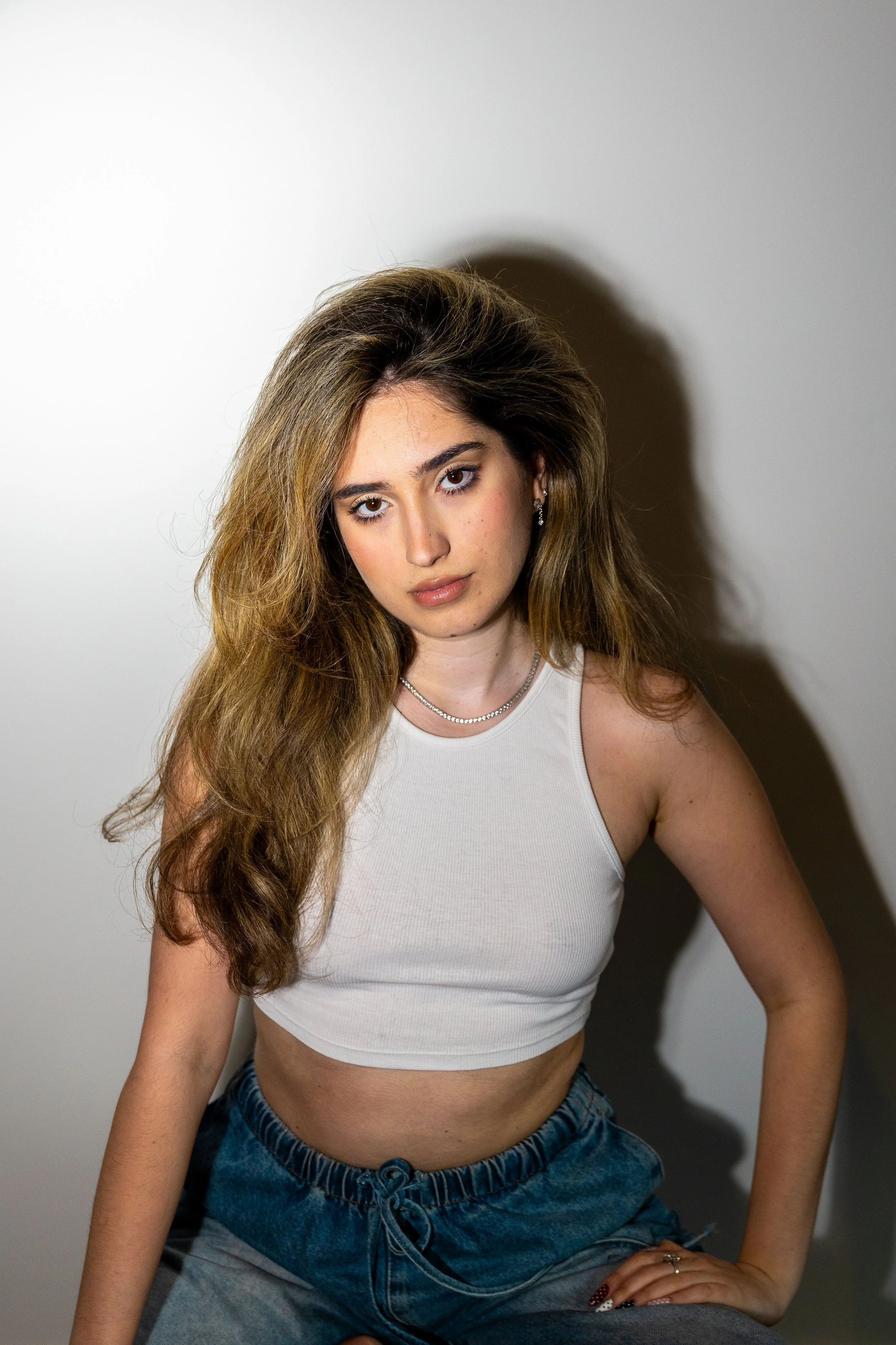 A young woman with long, wavy light brown hair, wearing a white sleeveless crop top, high-waisted blue denim shorts, and jewelry, sitting against a plain gray background with a shadow cast on the wall behind her.