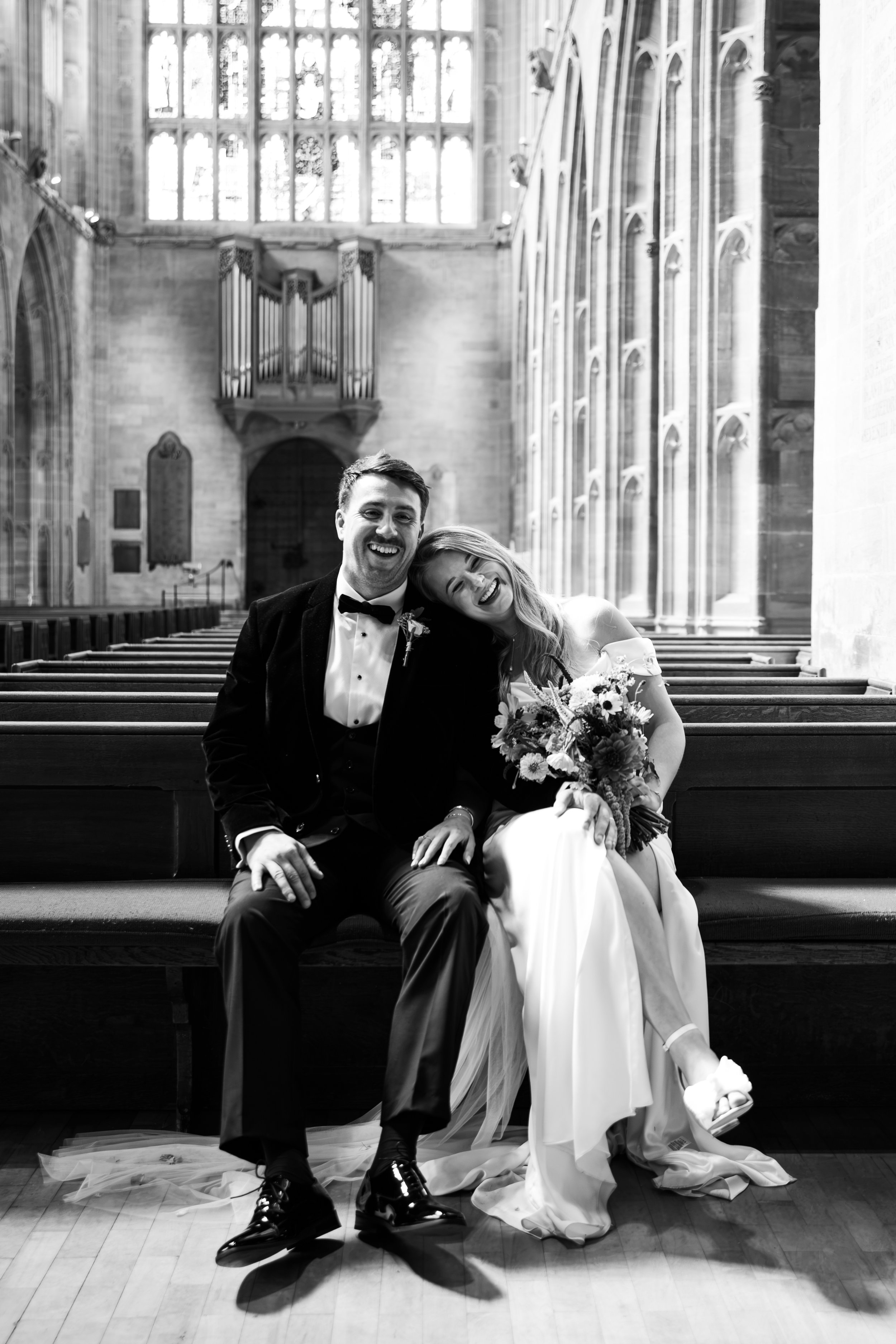 A happy bride and groom sitting on church pews inside a church. The bride is holding a bouquet, smiling with her head resting on the groom's shoulder. The groom is dressed in a tuxedo, smiling at the camera. The church interior features tall stained glass windows and an ornate pipe organ.