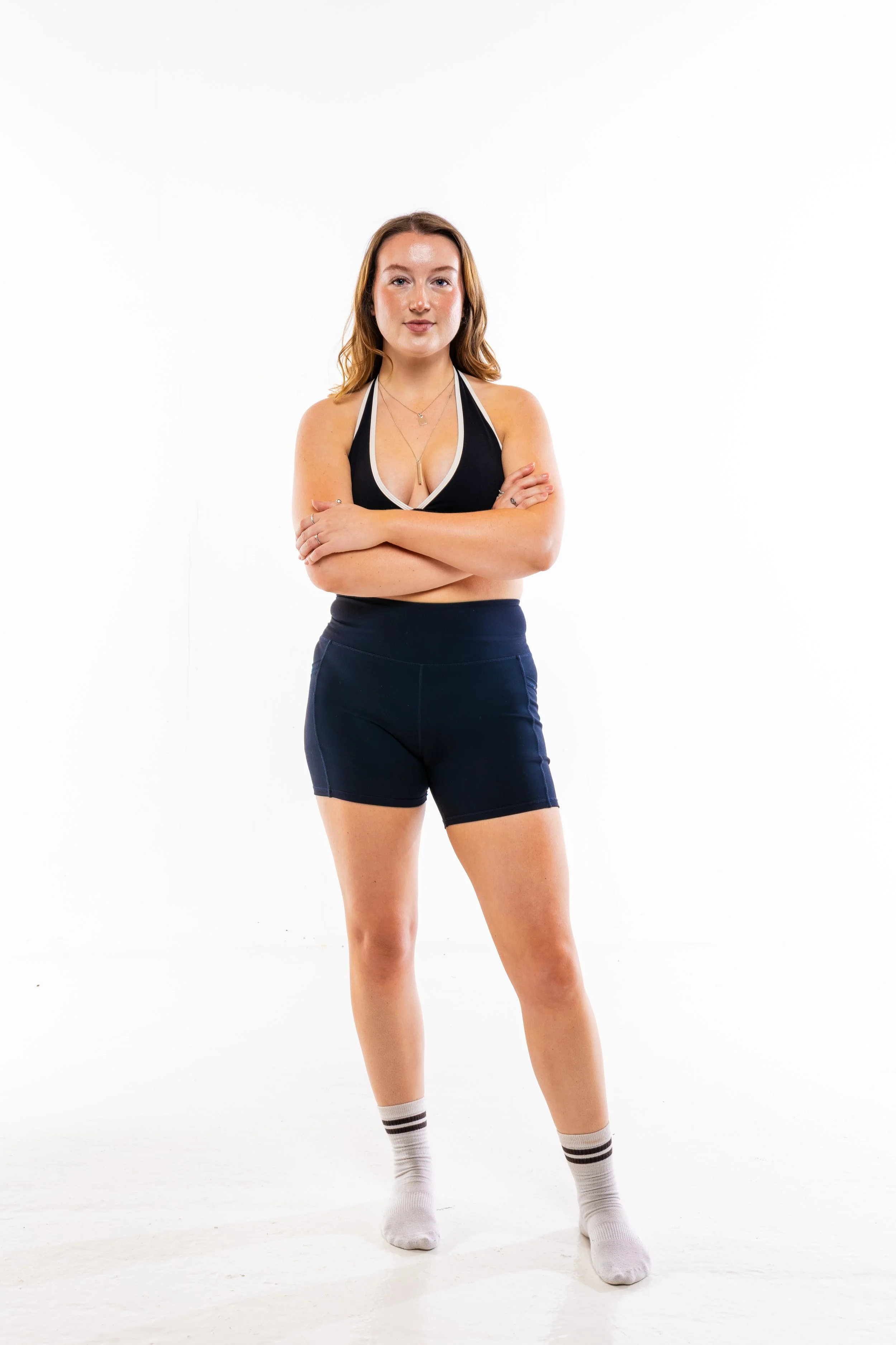 A young woman with light brown hair, standing with arms crossed, wearing a black sports bra, dark shorts, and white socks with black stripes, on a white background.