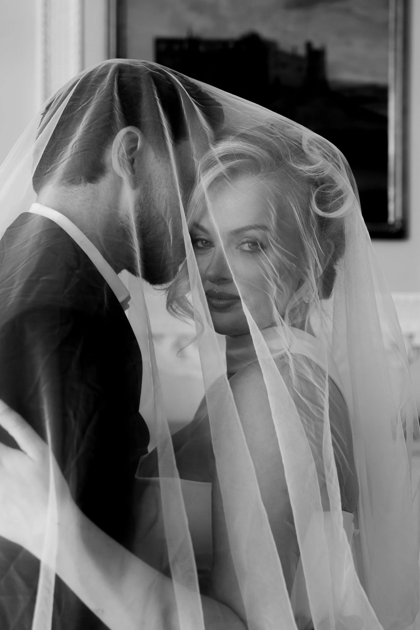 A black and white photo of a bride and groom close together, with the groom's face partly hidden and the bride's face fully visible, both under a sheer veil.