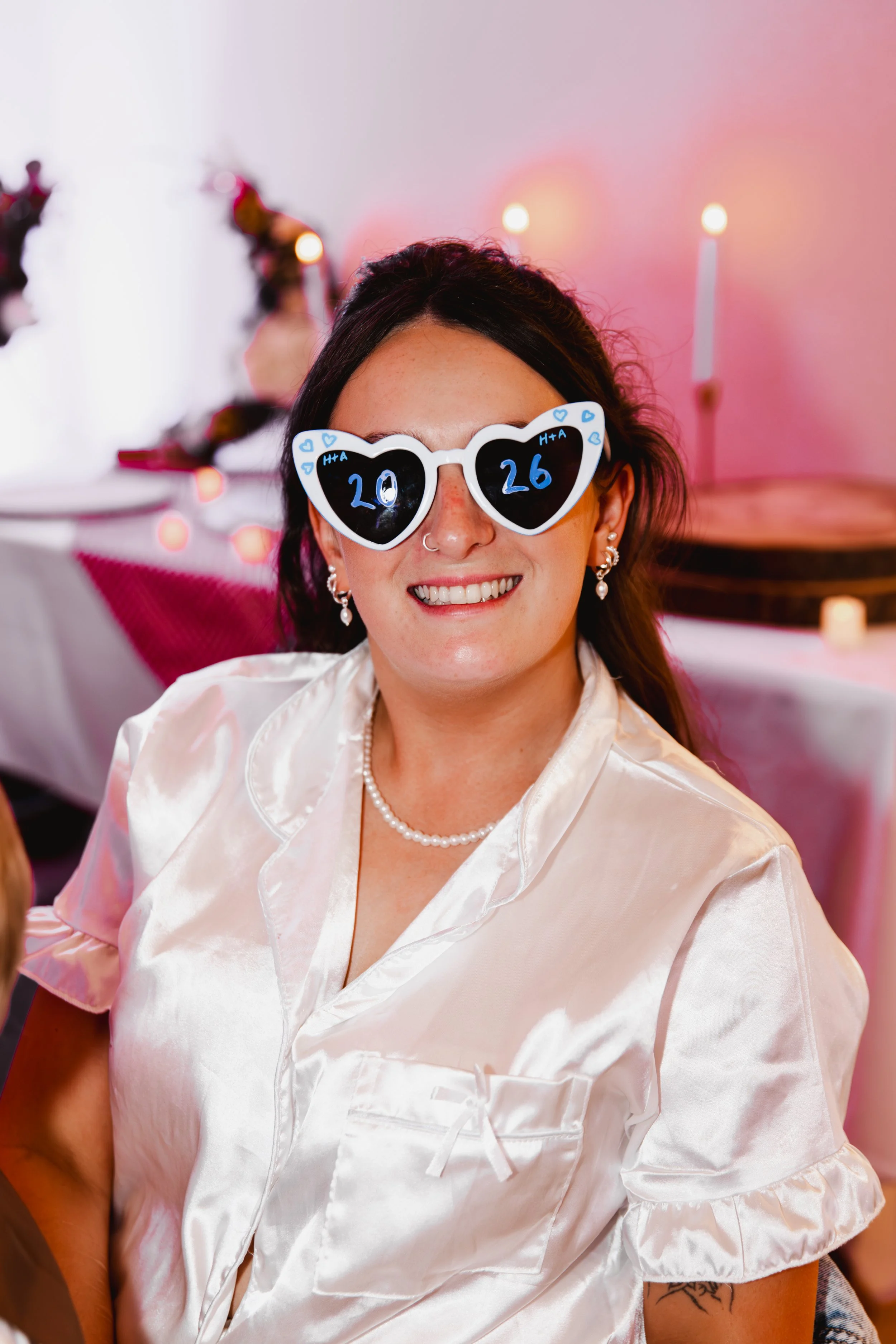 A woman with dark hair wearing heart-shaped sunglasses with the numbers 20 and 26 written on the lenses, smiling in a softly lit, pinkish background with blurred candles and flowers.