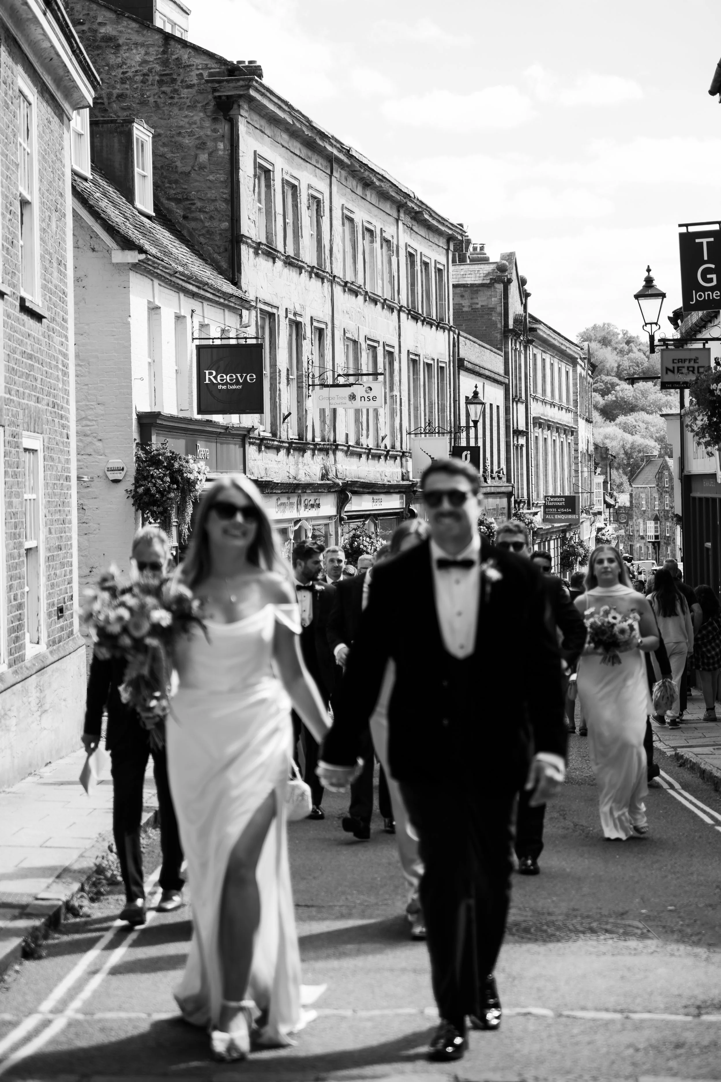 Black and white image of a street scene with several people dressed in formal wedding attire walking down the street, with buildings and store signs in the background.