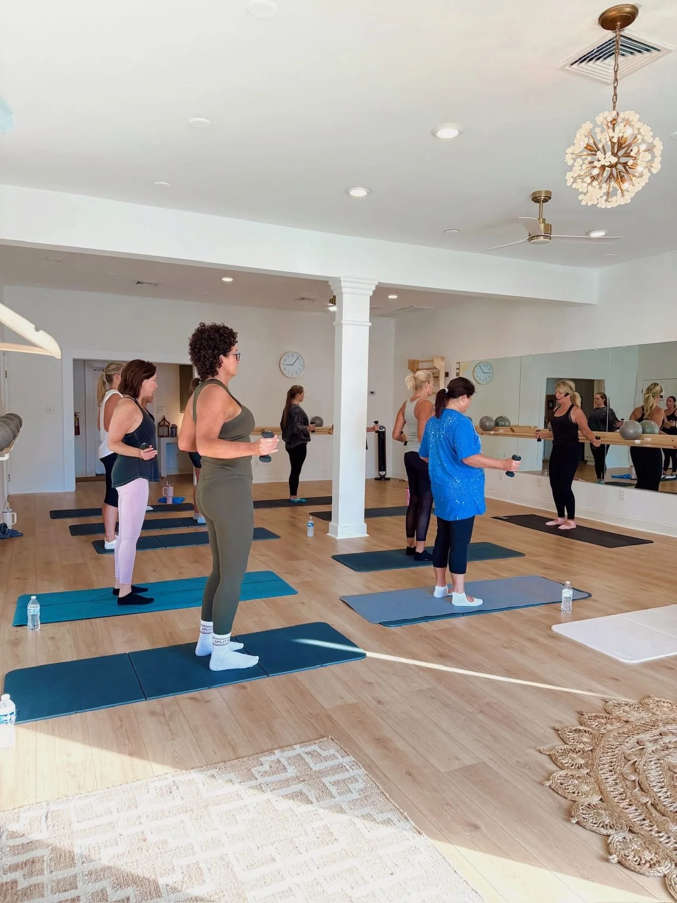 Women participating in a yoga or exercise class in a bright, spacious studio with large mirrors, mats, and props.