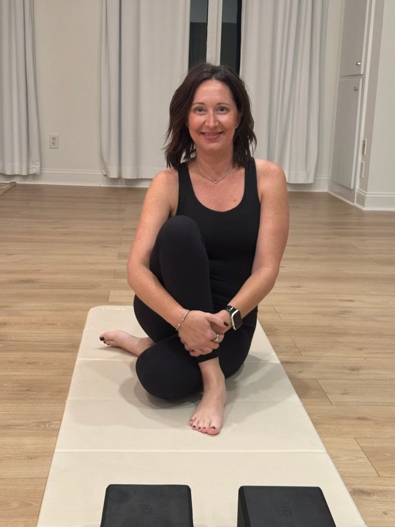 Woman with dark hair in workout clothes sitting on a yoga mat in a room with white curtains, with fitness blocks nearby.