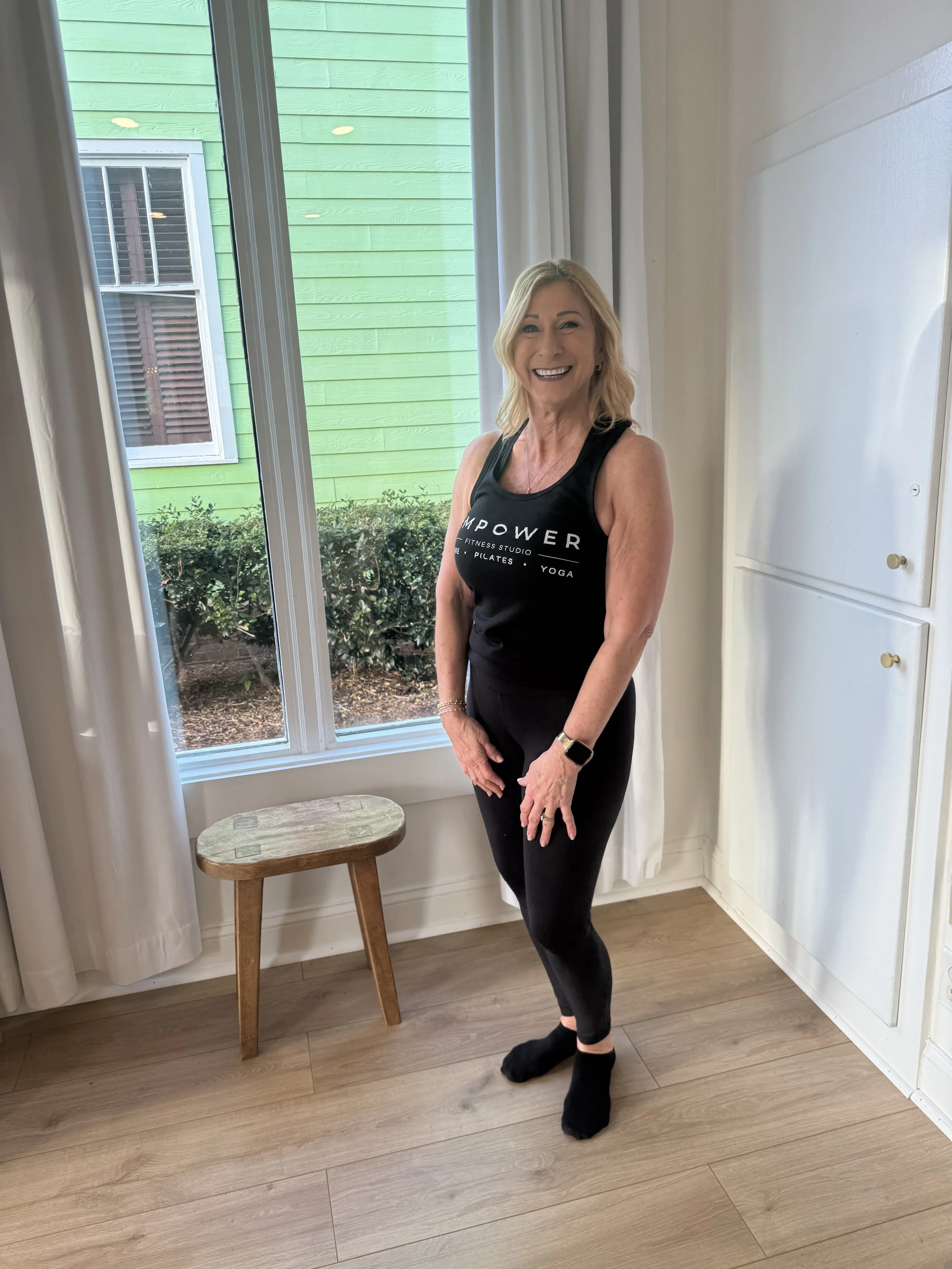 A woman with blonde hair smiling, wearing a black tank top and black leggings, standing indoors near a window with white curtains and a wooden stool, with a green exterior wall outside.
