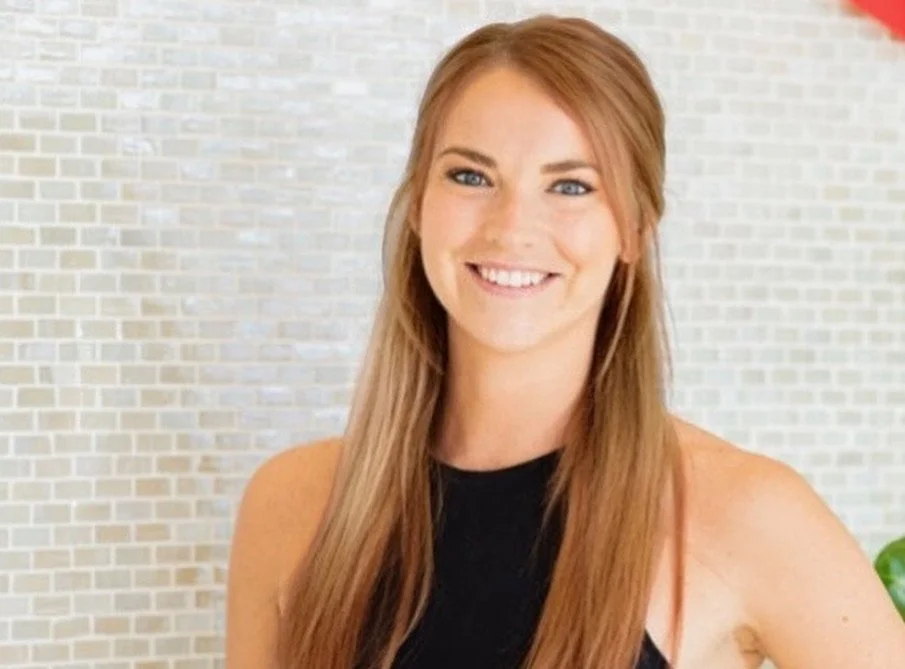 A woman with long red hair smiling at the camera, wearing a black sleeveless top, standing in front of a textured wall.