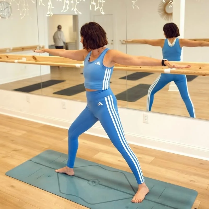 Woman in blue athletic wear practicing ballet or yoga with a wooden barre in front of a wall mirror in a dance studio.