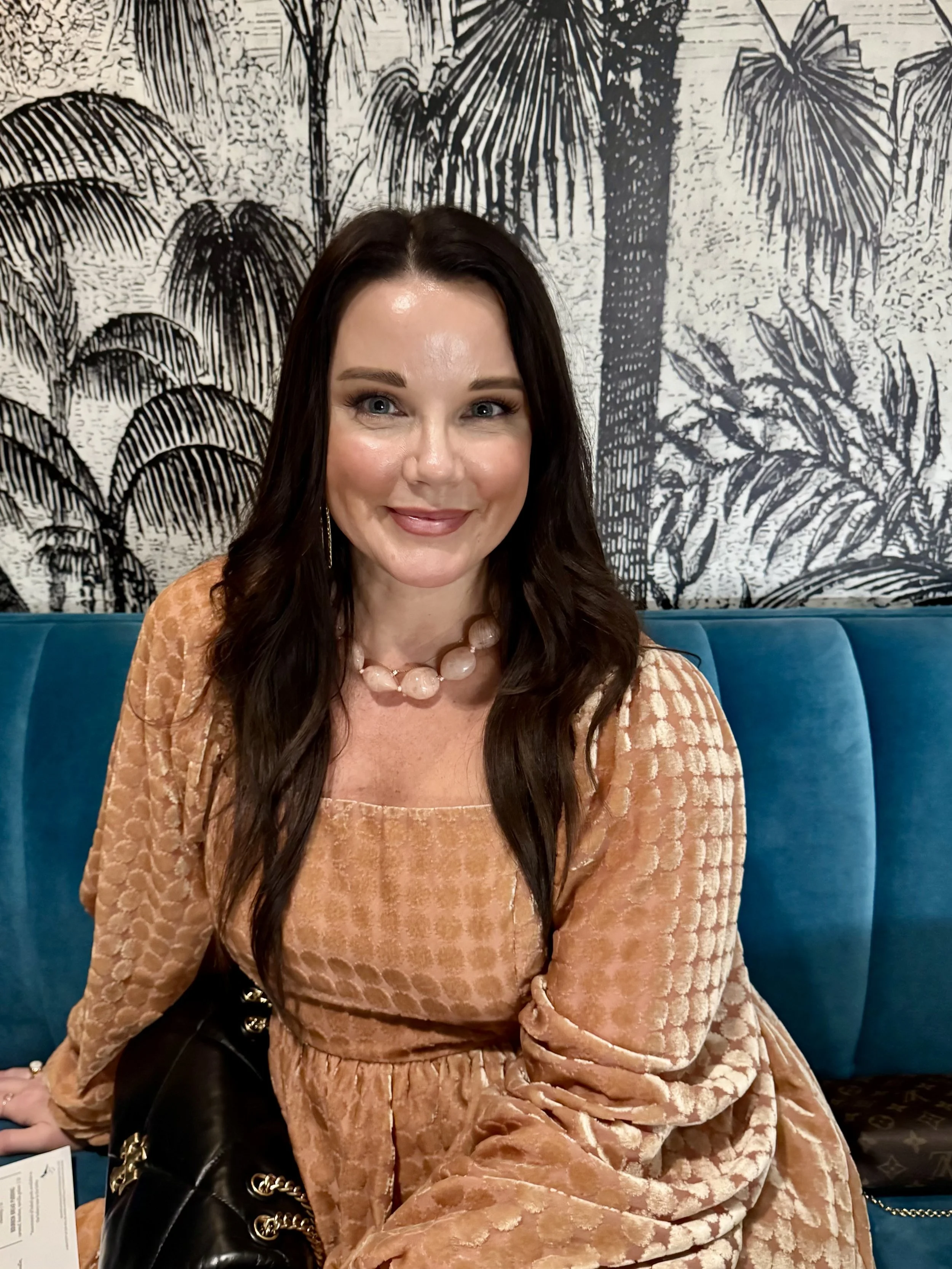 A woman with long dark hair, light skin, and blue eyes, smiling and sitting on a blue couch in front of a black and white botanical-themed wall mural, wearing a peach-colored textured dress and a large bead necklace.