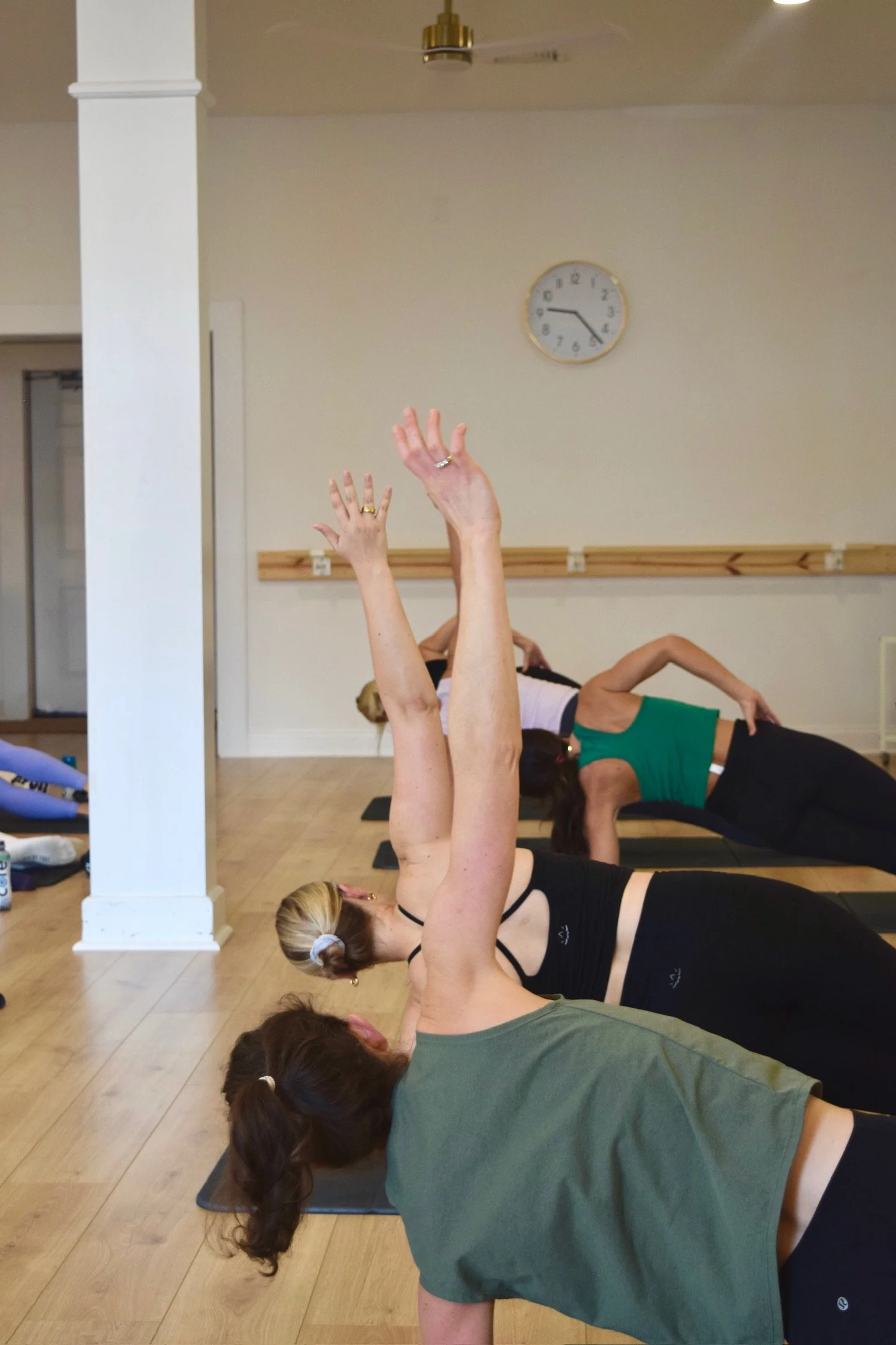 Group of women practicing yoga in a class, lying on mats with arms raised overhead in a side stretch pose in a studio with a clock on the wall.