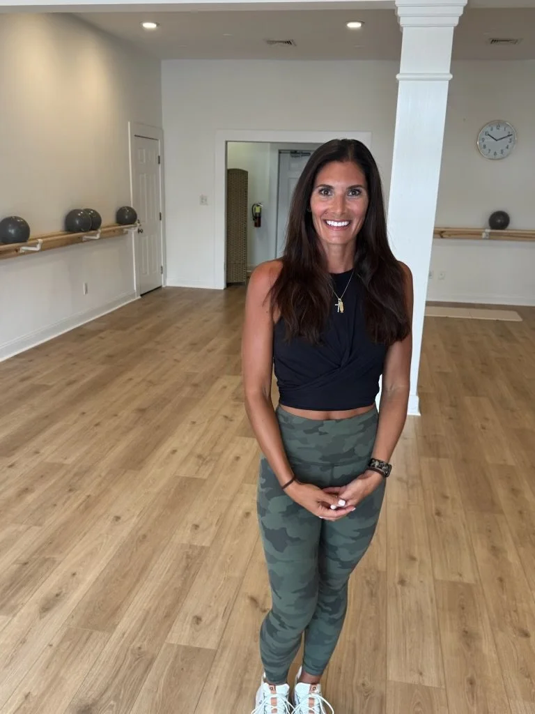 A woman with long dark hair wearing a black sleeveless crop top, camouflage leggings, and white sneakers, smiling standing in a dance studio with wooden floors and wall-mounted ballet barres.