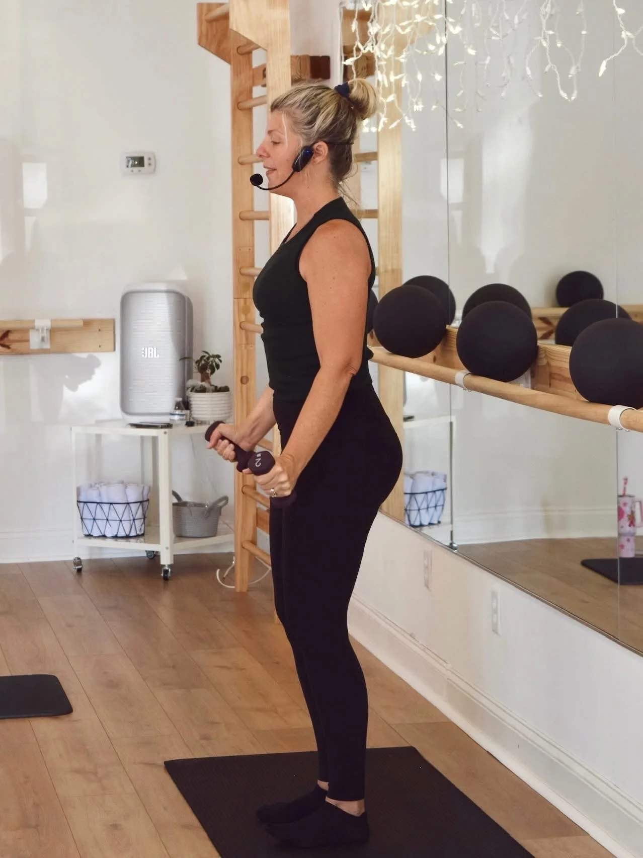 A woman with a headset holding a dumbbell in a fitness studio with black exercise balls on a shelf and a mirror.