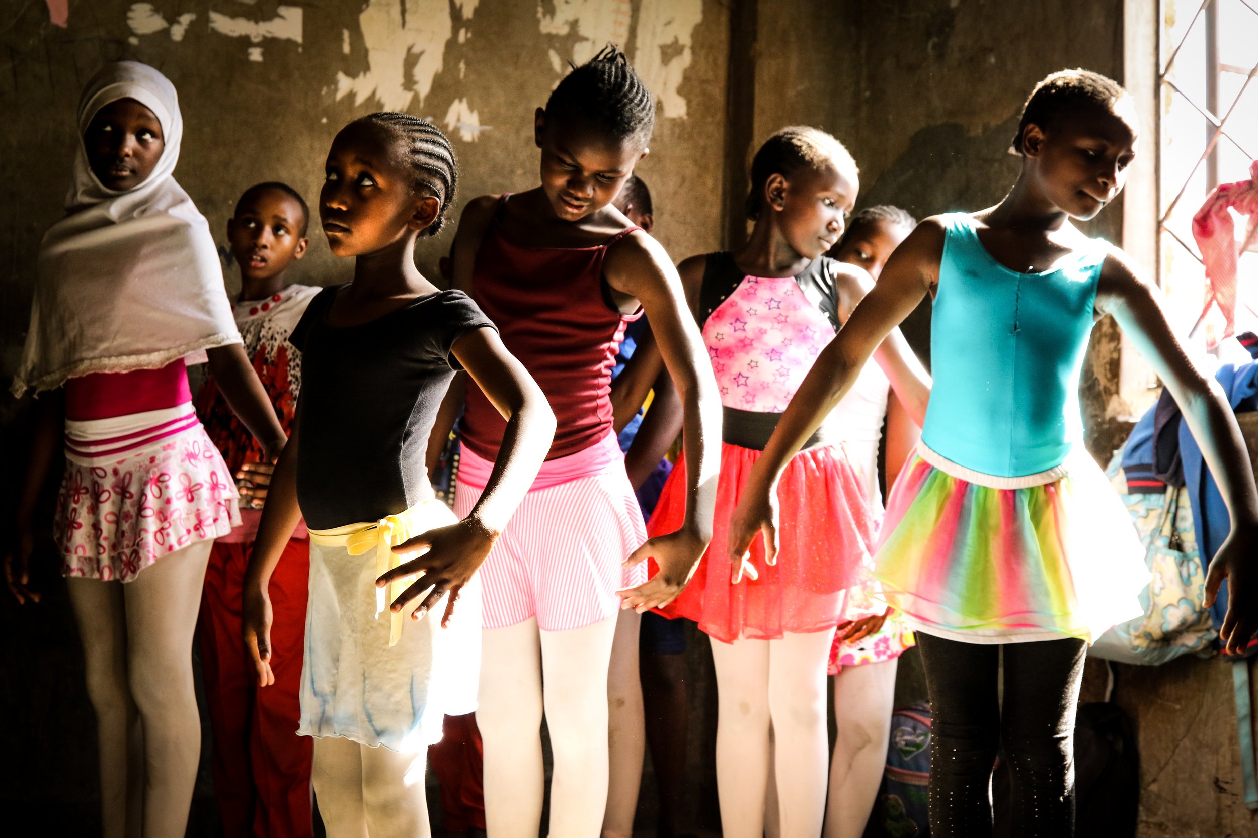 Group of young girls dressed in colorful dance costumes practicing ballet in a dimly lit room.