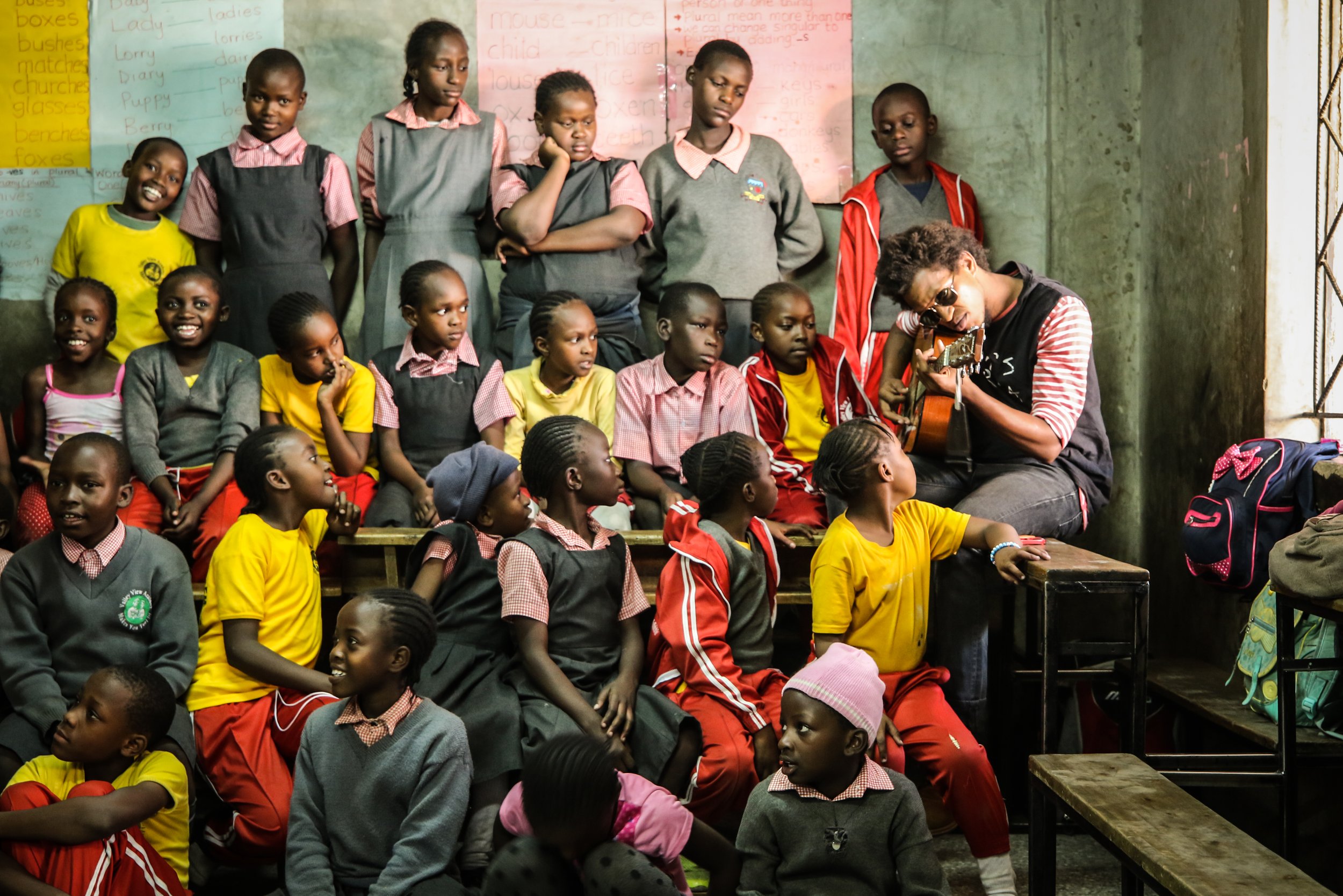 A group of school children sitting and standing inside a classroom, watching a person playing guitar.