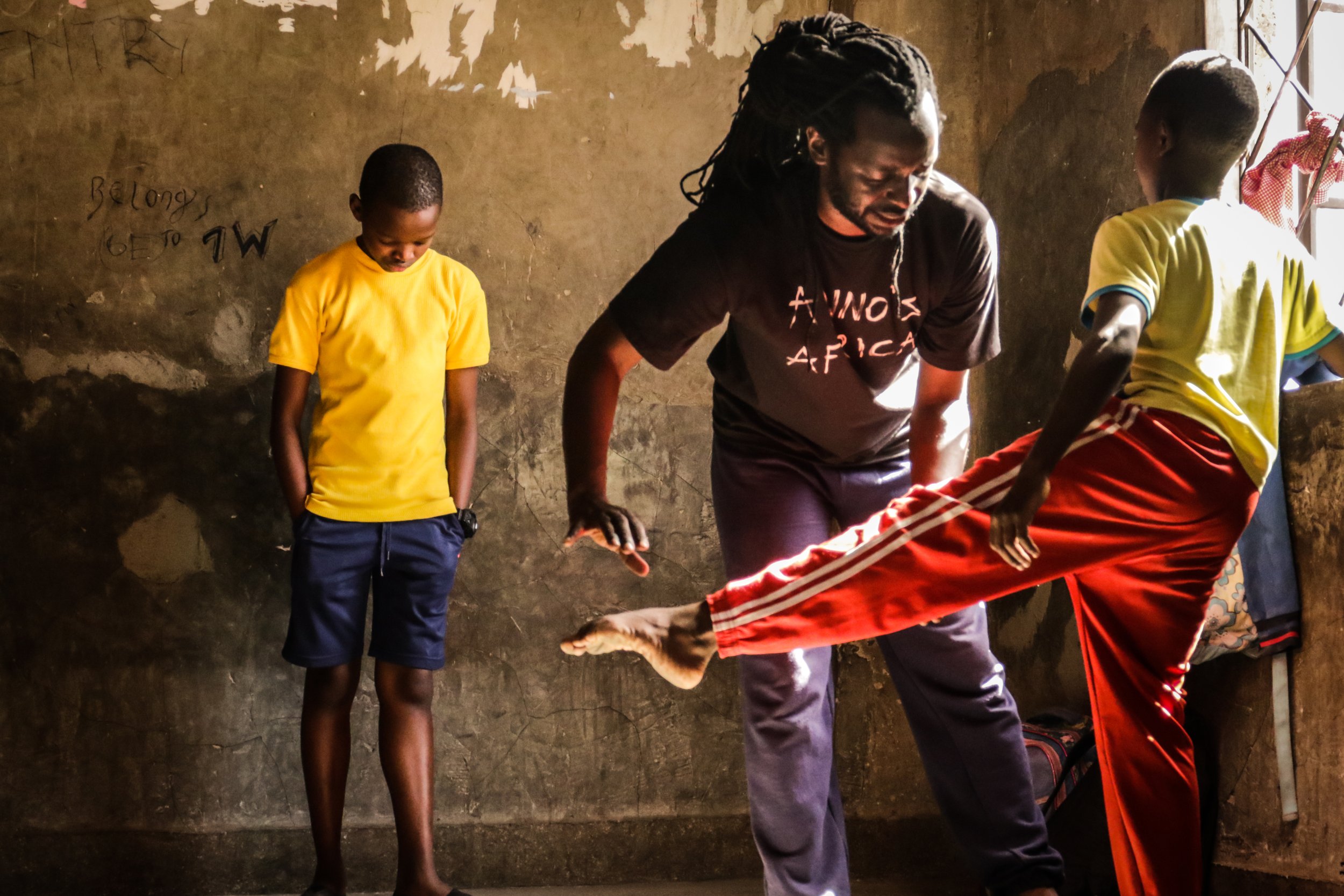 A man with long dreadlocks and a black t-shirt practicing martial arts with a boy wearing a yellow t-shirt, while two other boys in yellow and red pants watch in a room with a worn wall and sunlight coming through a window.