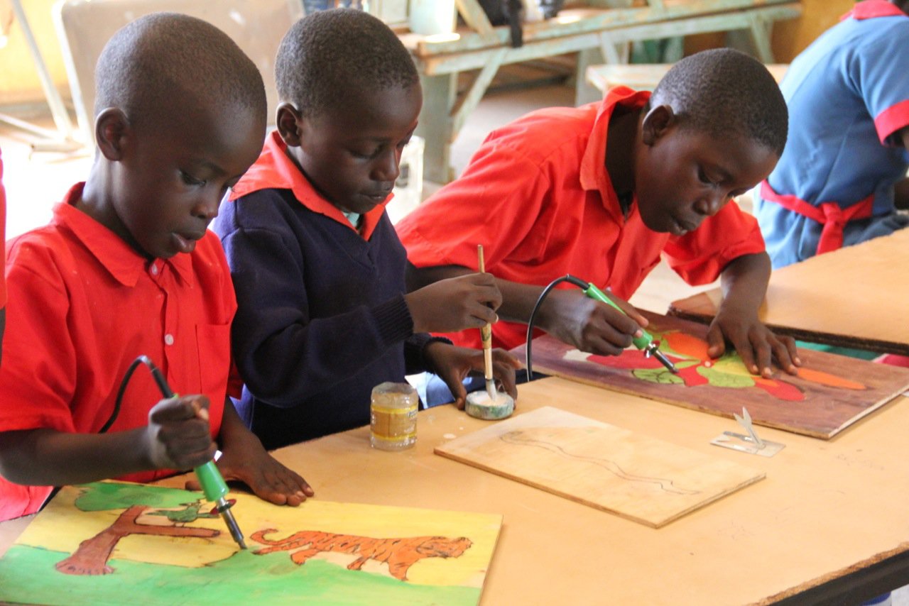 Three boys working on colorful paintings of trees and tigers with soldering tools in a classroom setting.