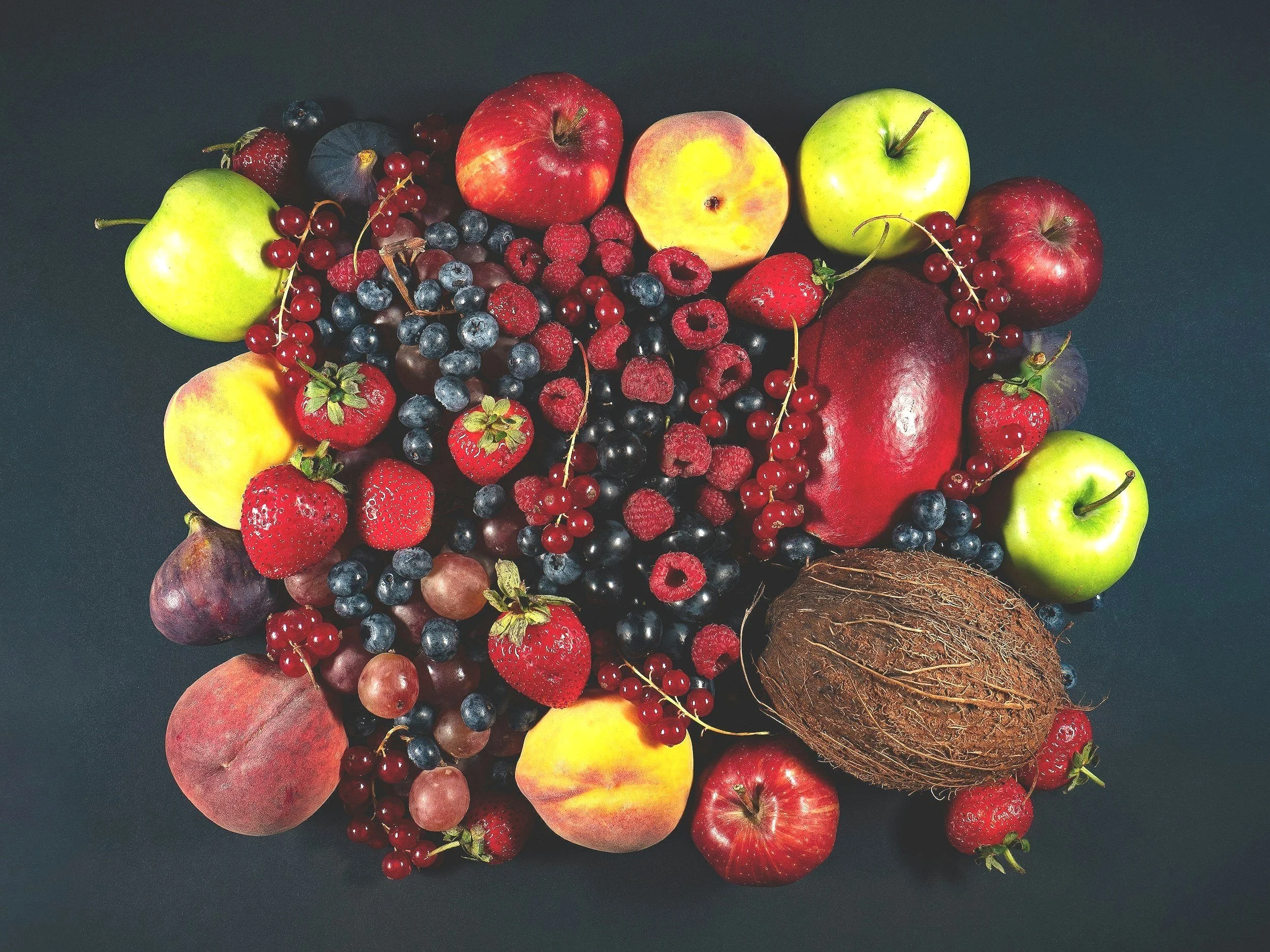 A variety of fresh fruits including apples, peaches, strawberries, blueberries, raspberries, figs, currants, grapes, and a coconut arranged on a dark background.