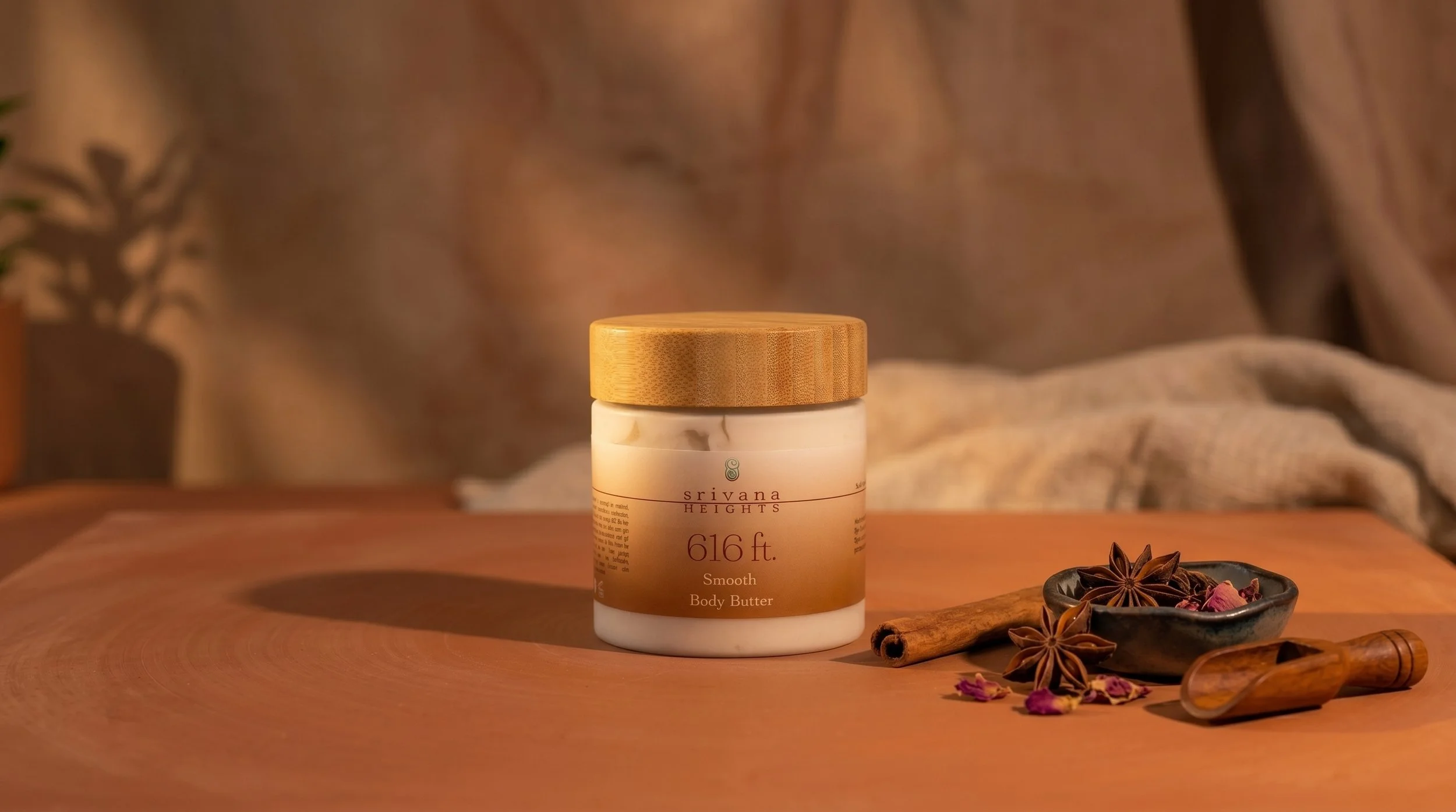 A jar of Srivana Heights body butter on a wooden surface, with star anise and cinnamon sticks next to it, and a bowl of dried flower petals nearby.