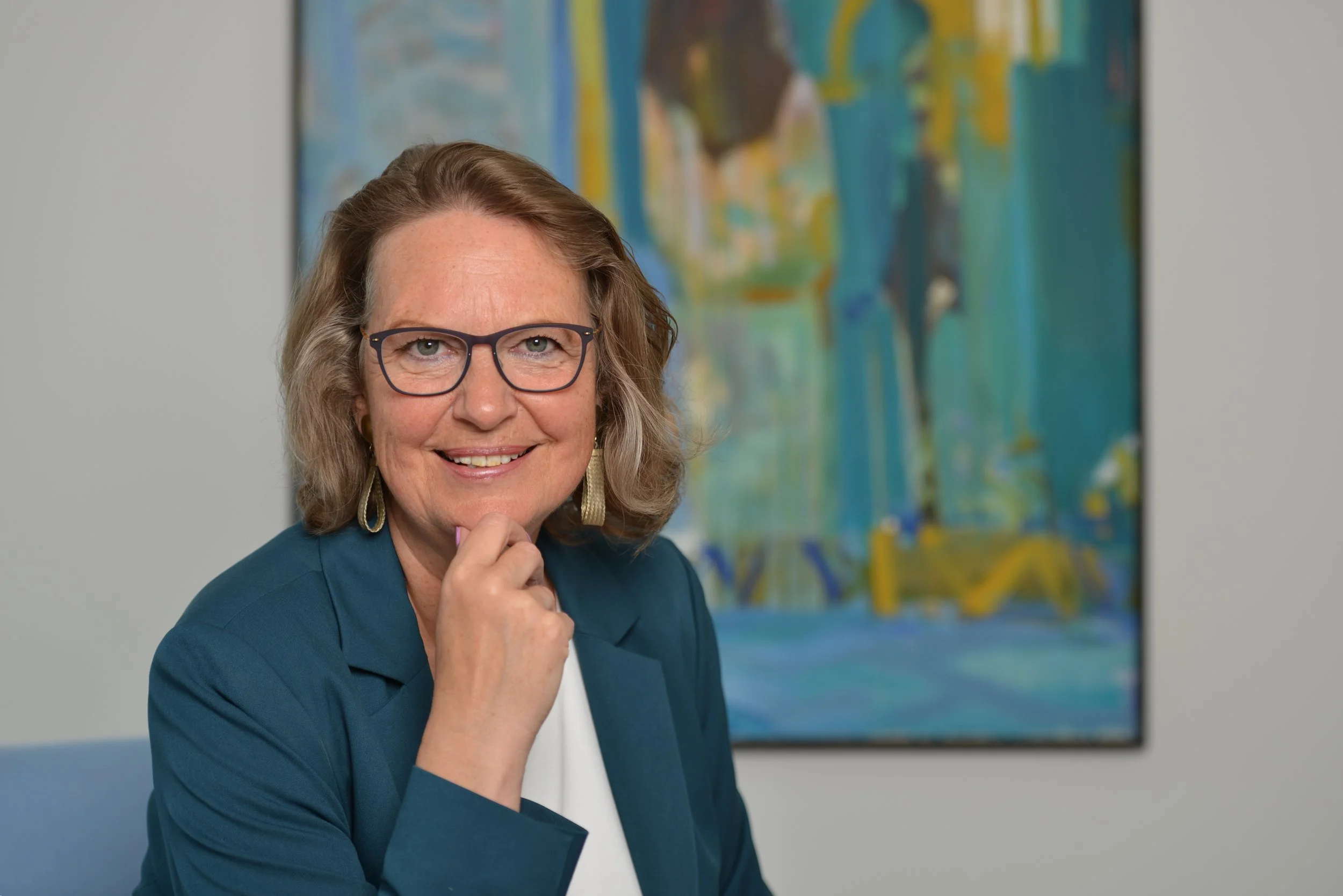 Porträt einer lächelnden älteren Frau mit Brille und Locken, die in einem blauen Blazer sitzt vor einem abstrakten Gemälde.