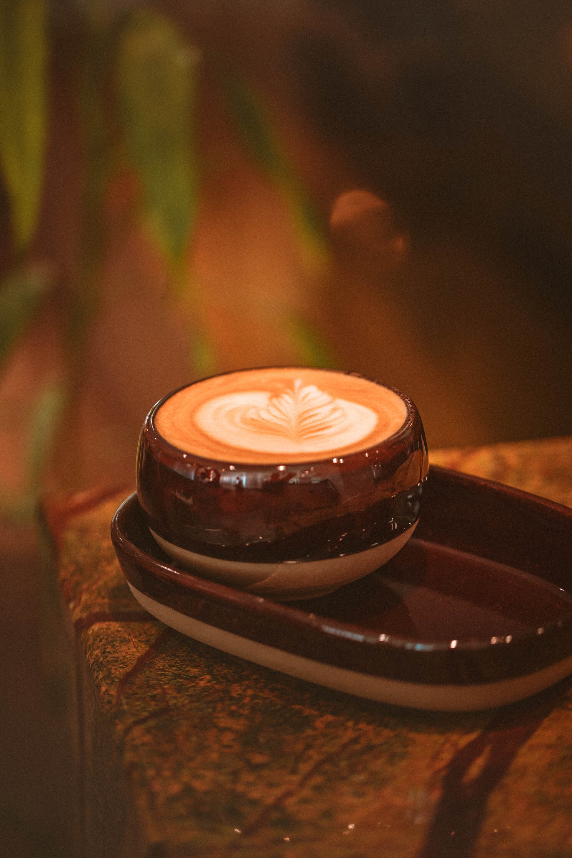 Un cappuccino avec un pour de latte art dans une tasse en céramique sur une surface en bois, avec un décor flou en arrière-plan.