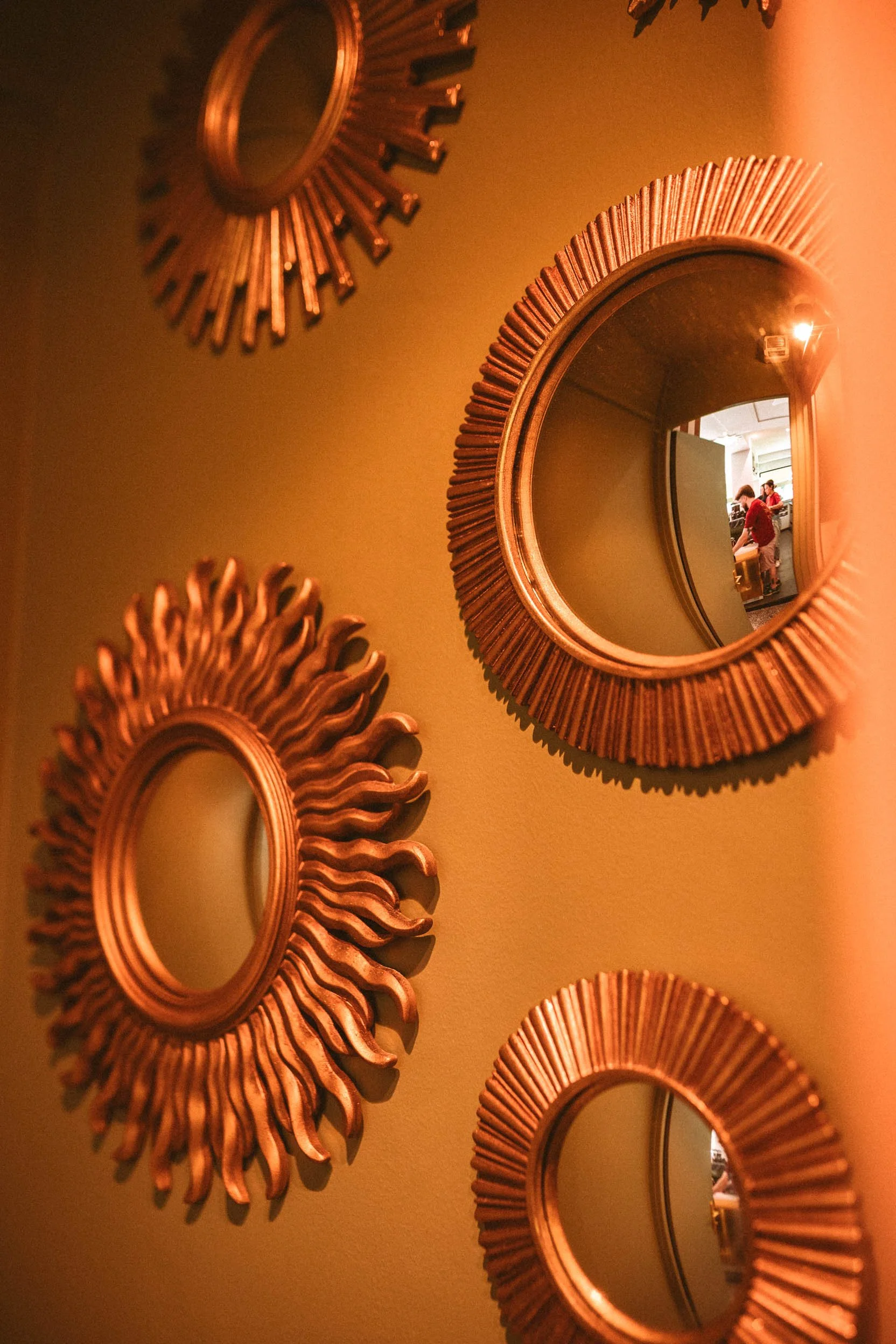 Une collection de miroirs ronds à cadre en métal doré avec des rayures, accrochés sur un mur avec un éclairage chaud. La réflexion dans un miroir montre deux personnes, dont une portant un vêtement rouge, dans une pièce intérieure.