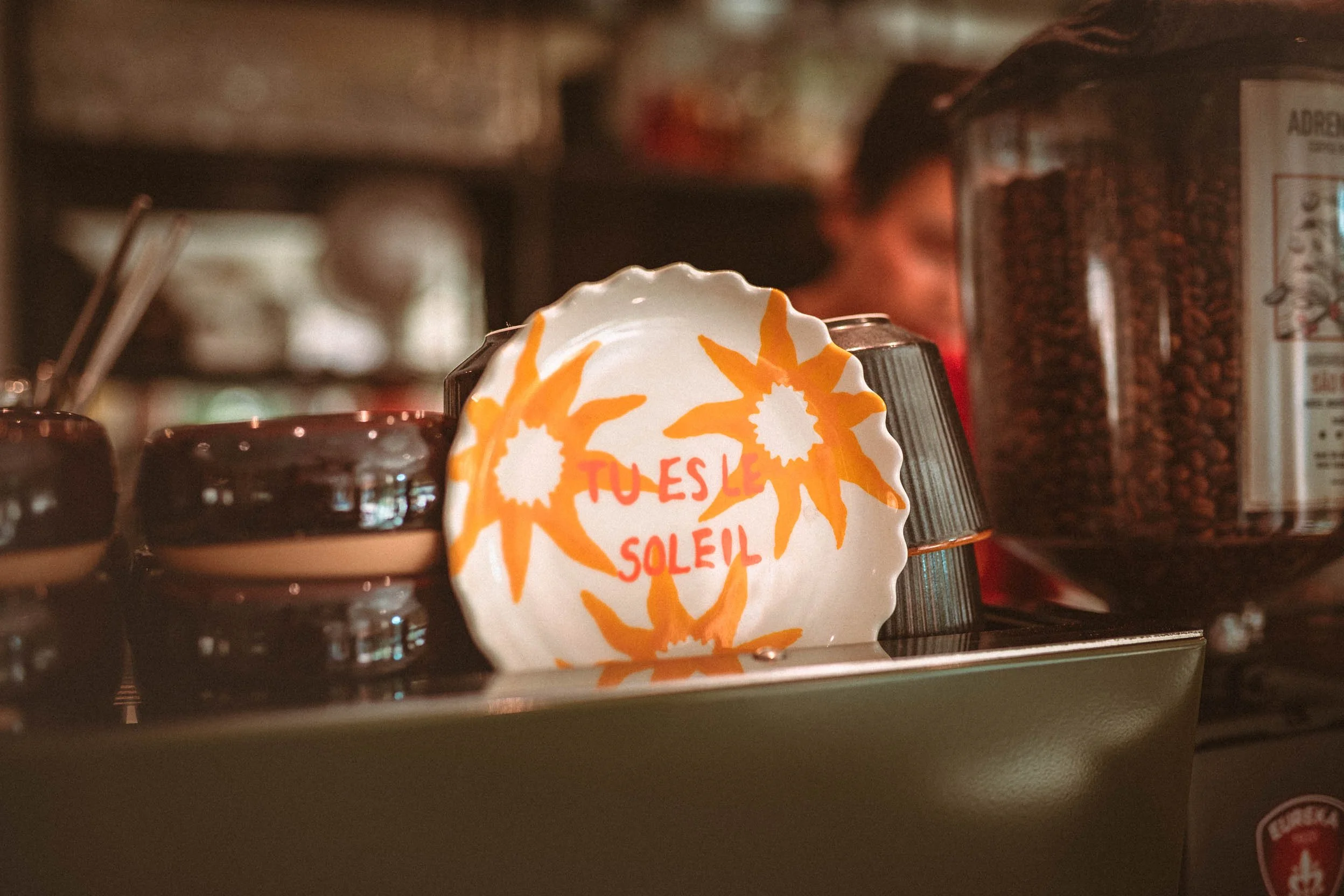 Assiette en céramique blanche avec des fleurs orange et le texte 'Tu es le soleil' écrit dessus, placée parmi des cafetières et des contenants de café, avec en arrière-plan une personne floue.