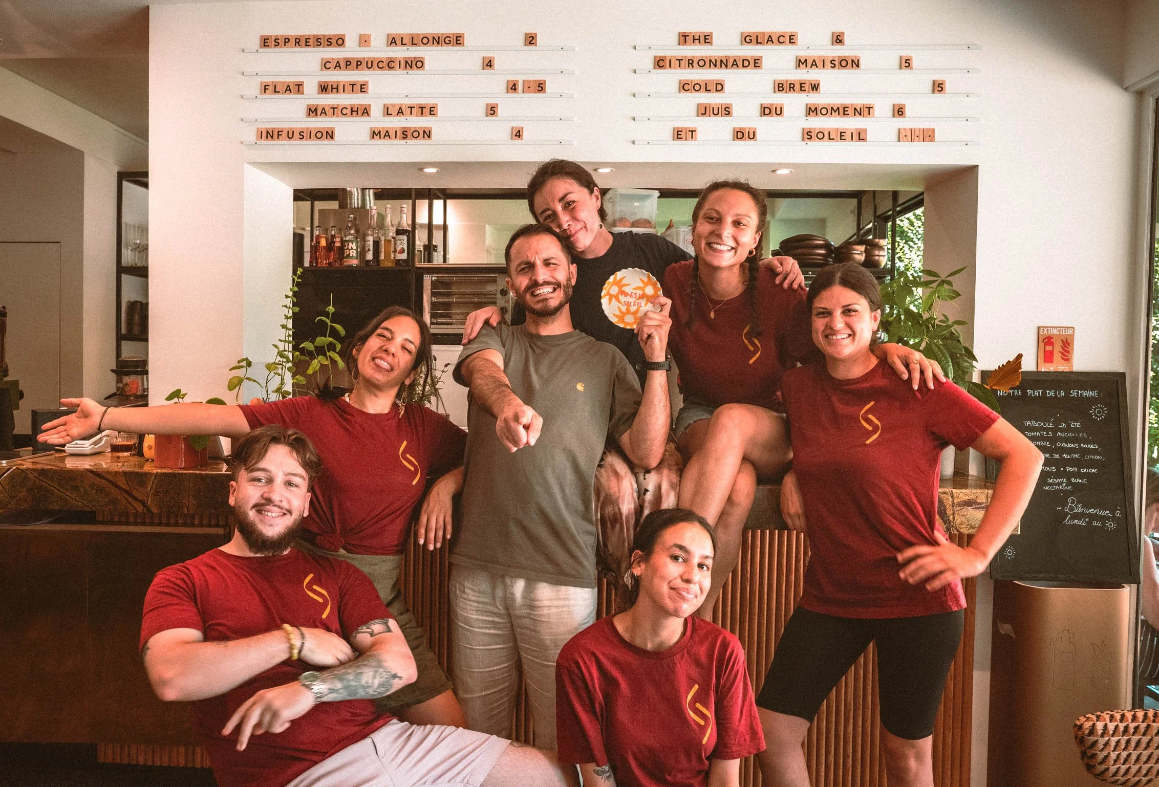 Groupe de sept personnes souriantes, posant dans un café ou restaurant, portant des T-shirts rouges avec un logo jaune, certains avec des bras autour des autres, à l'intérieur d'un établissement avec une ardoise de menu en arrière-plan.