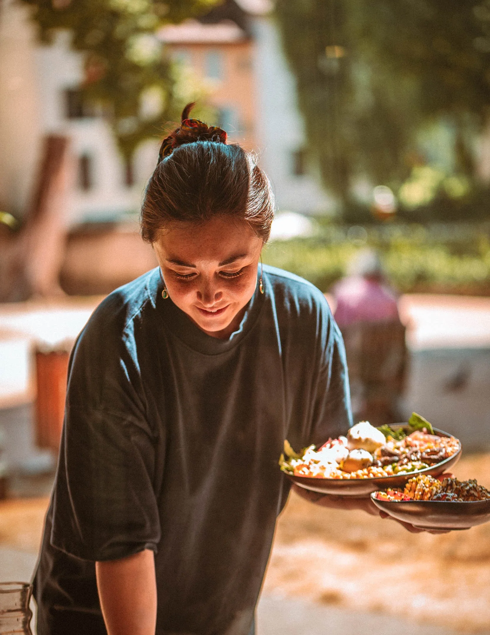 Une femme souriante partageant un repas en plein air, tenant deux assiettes remplies de nourriture.