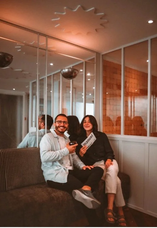 Deux jeunes adultes assis sur un canapé dans un intérieur moderne, souriant vers la caméra, avec un homme tenant un verre et une femme tenant une carte ou un signe, dans un environnement chaleureux avec des miroirs et un mur en briques en arrière-plan.