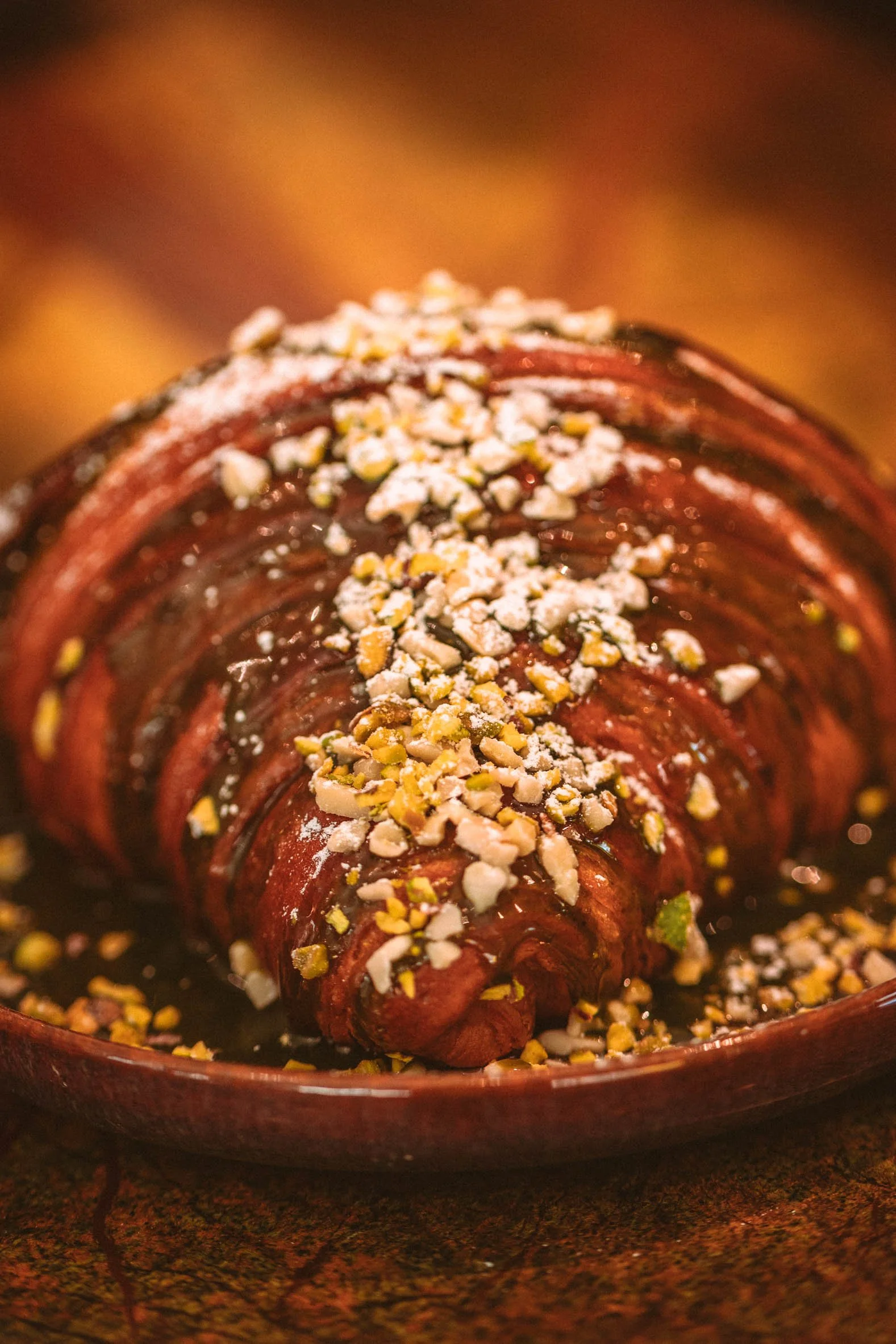 Un croissant chocolaté nappé de glaçage au chocolat et parsemé de noix hachées, présenté dans une coupelle en terre cuite.