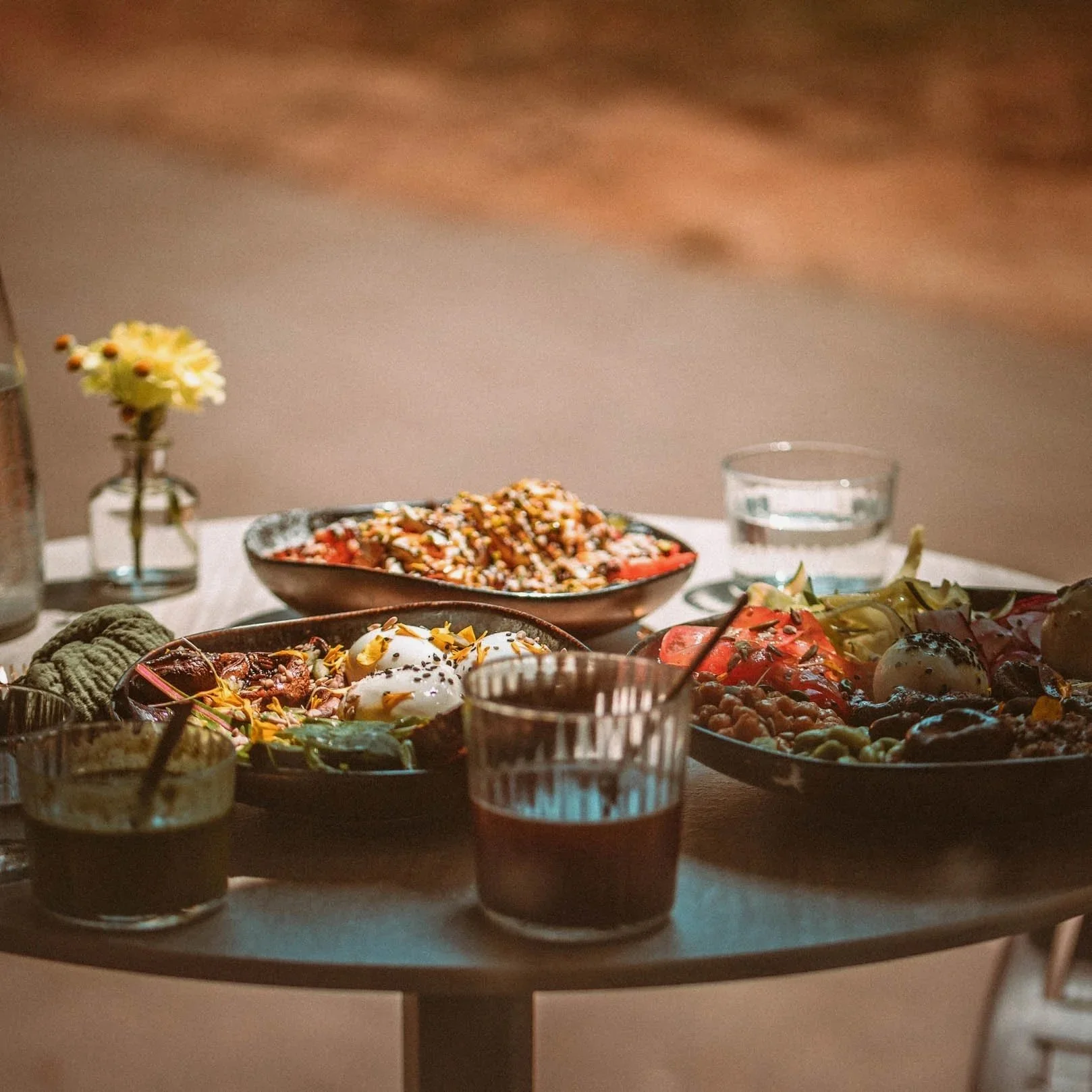Un repas avec plusieurs plats sur une table, comprenant des salades, des œufs pochés, des boissons et une petite fleur dans un vase.