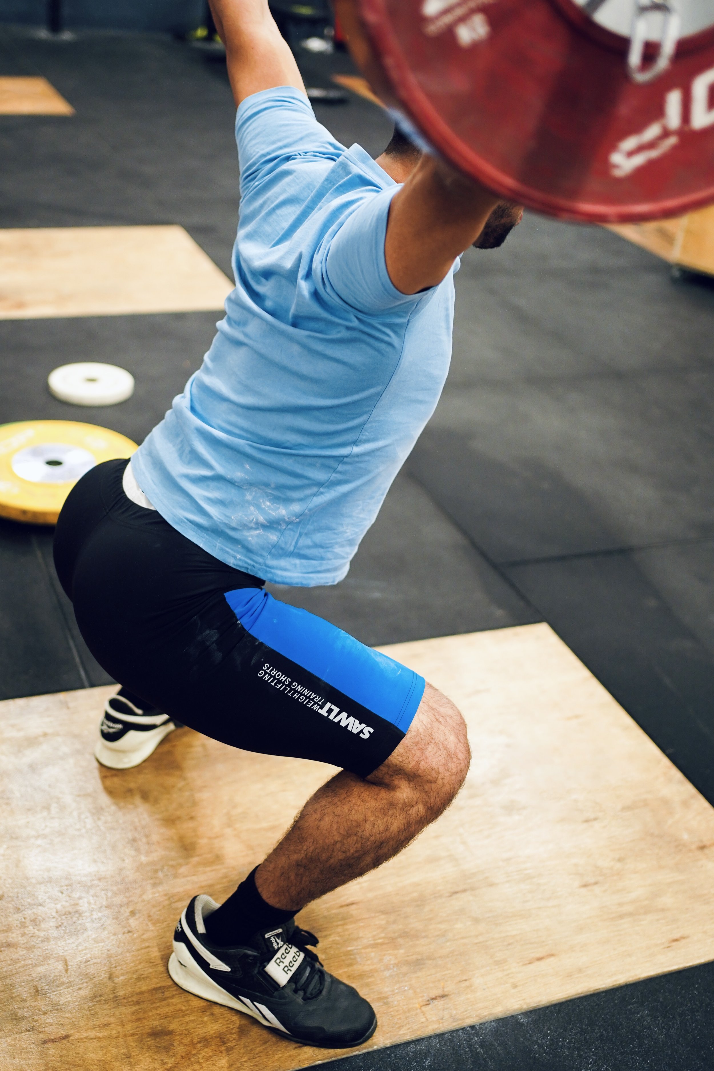 A man performing a snatch with a barbell loaded with red weight plates in a gym, wearing a light blue t-shirt, black shorts with blue accents, and black athletic shoes.