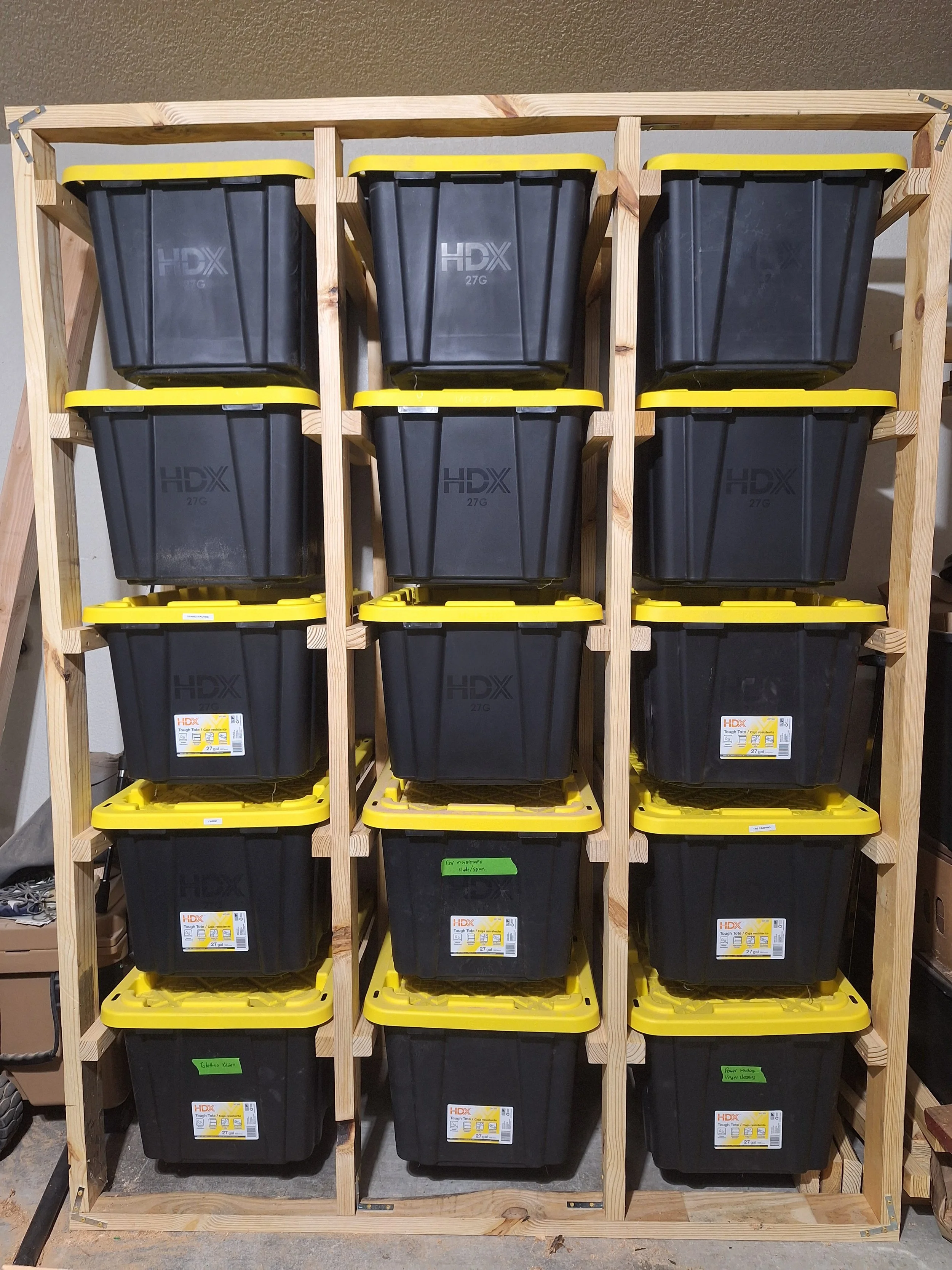 A wooden shelving unit holding multiple black storage bins with yellow lids, arranged in four columns and four rows, labeled with brand and contents.