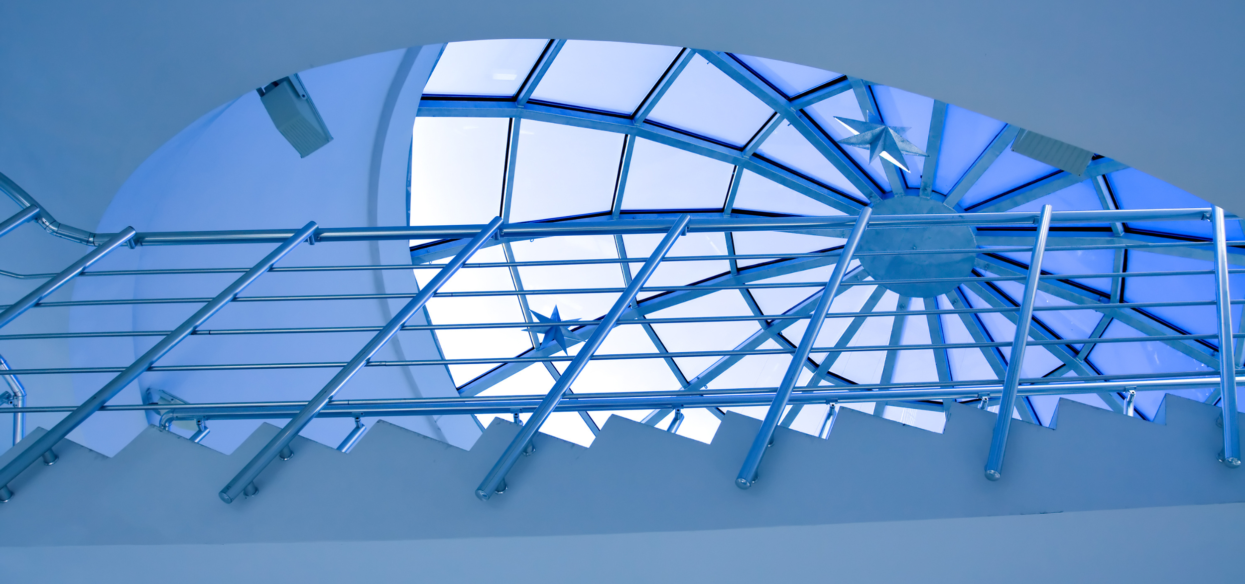 Looking up at a glass ceiling with a blue tint, supported by metal beams and decorated with star-shaped ornaments.