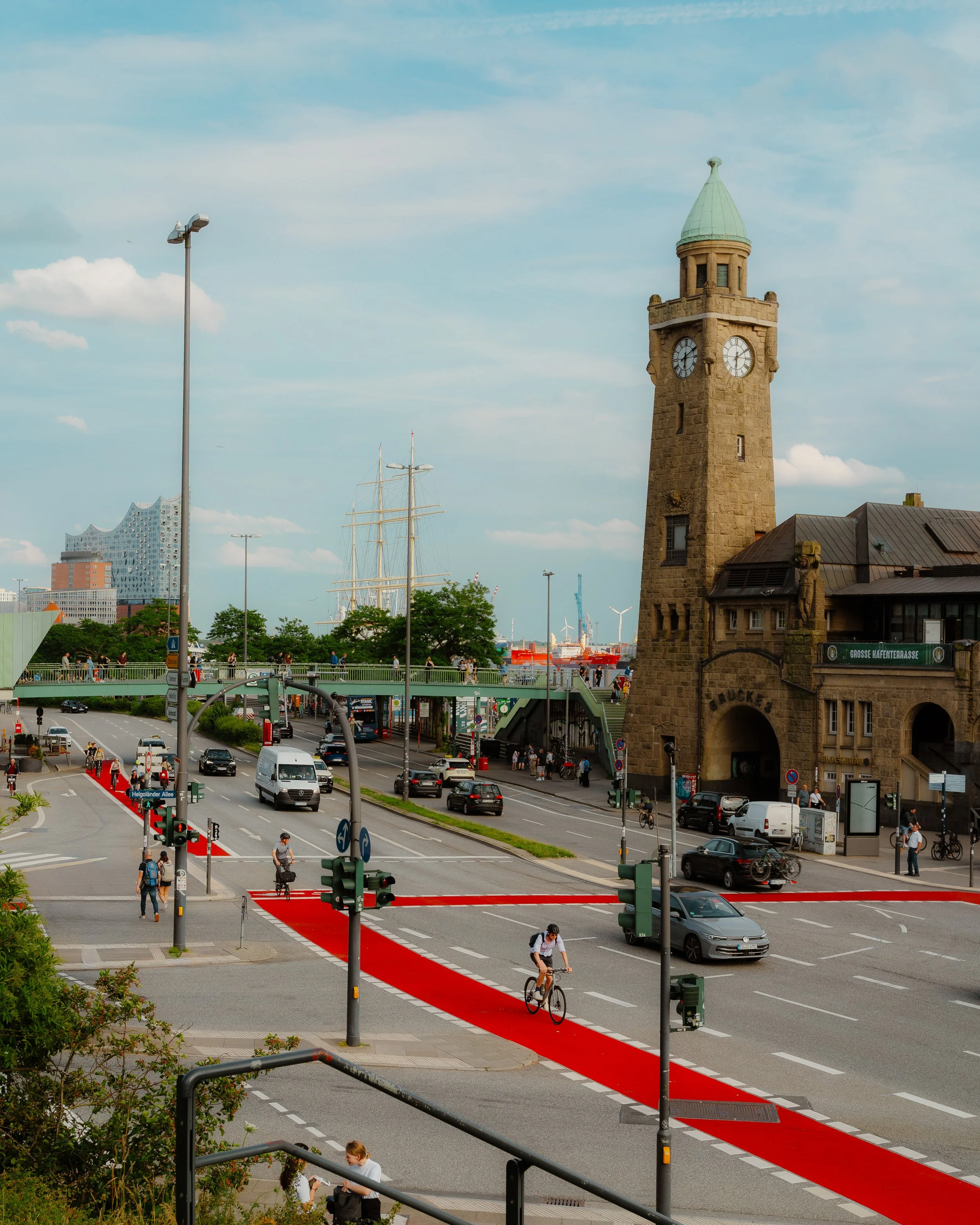 City street scene with a historic clock tower, cyclists, cars, pedestrians, and a red bike lane, with a modern building and ships in the background.