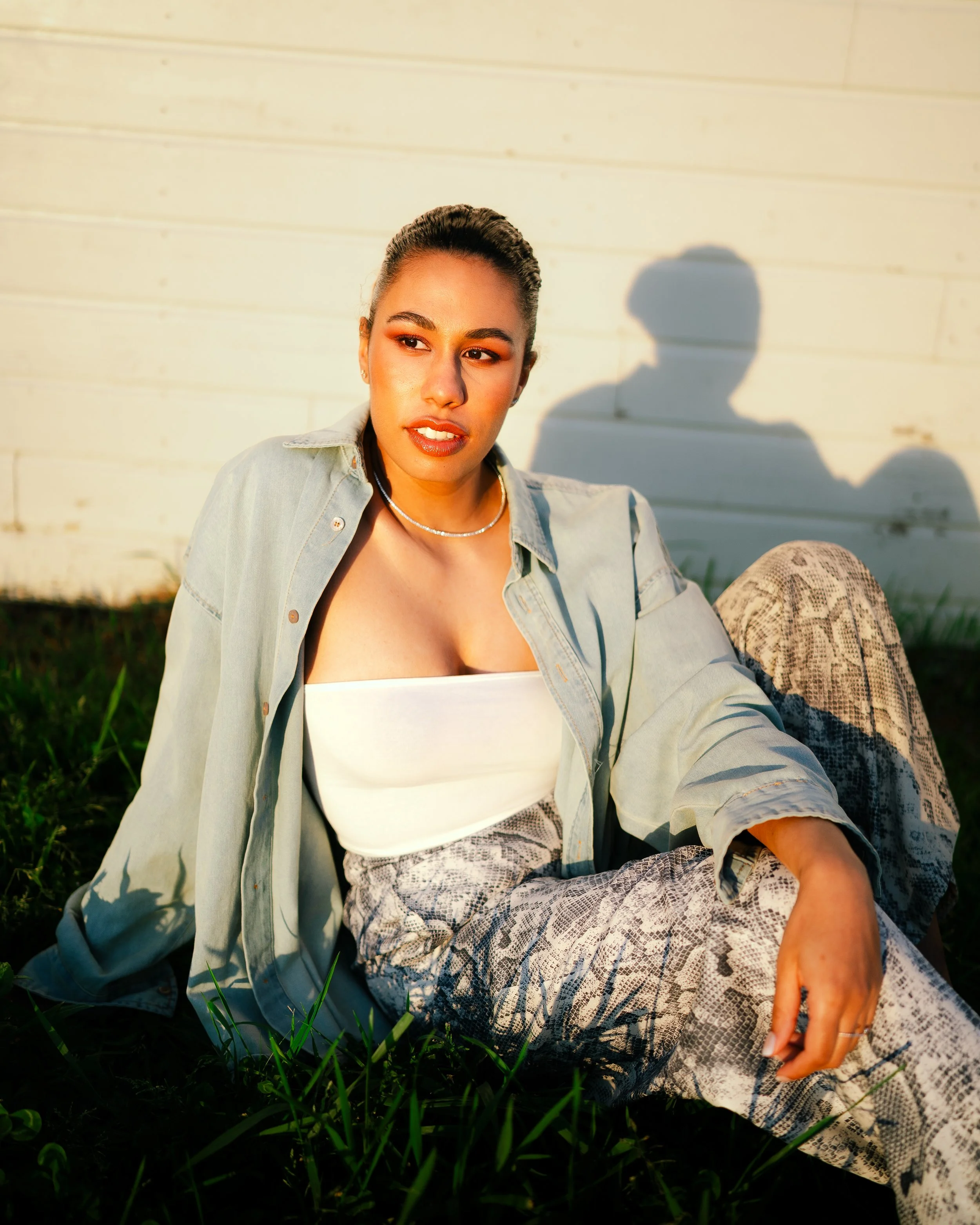 A woman sitting on the grass with a white wooden wall in the background, illuminated by warm sunlight, casting a shadow of her head and shoulder on the wall.