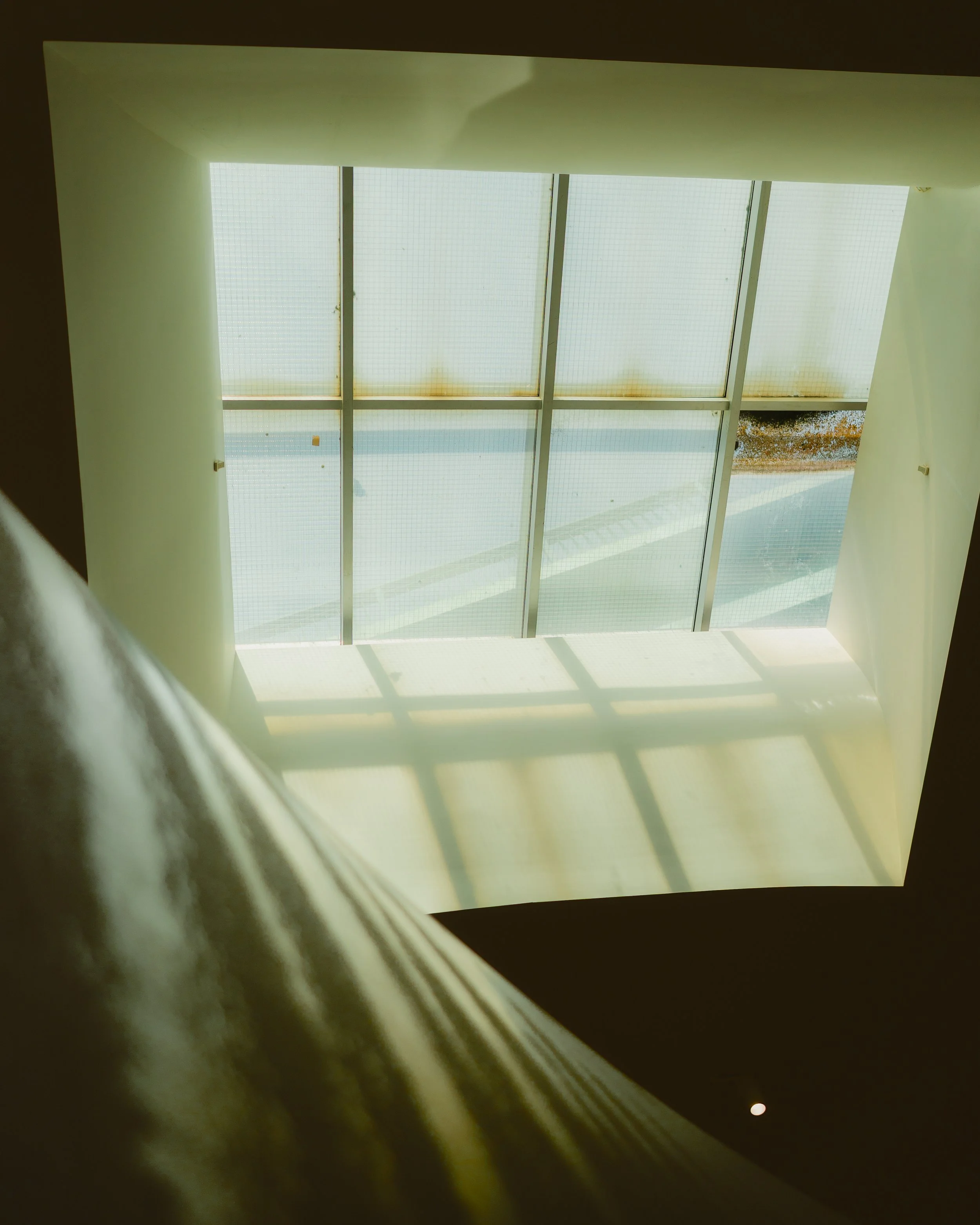 Looking up through a large skylight window with frosted and clear glass panels, sunlight casting shadows on the walls.