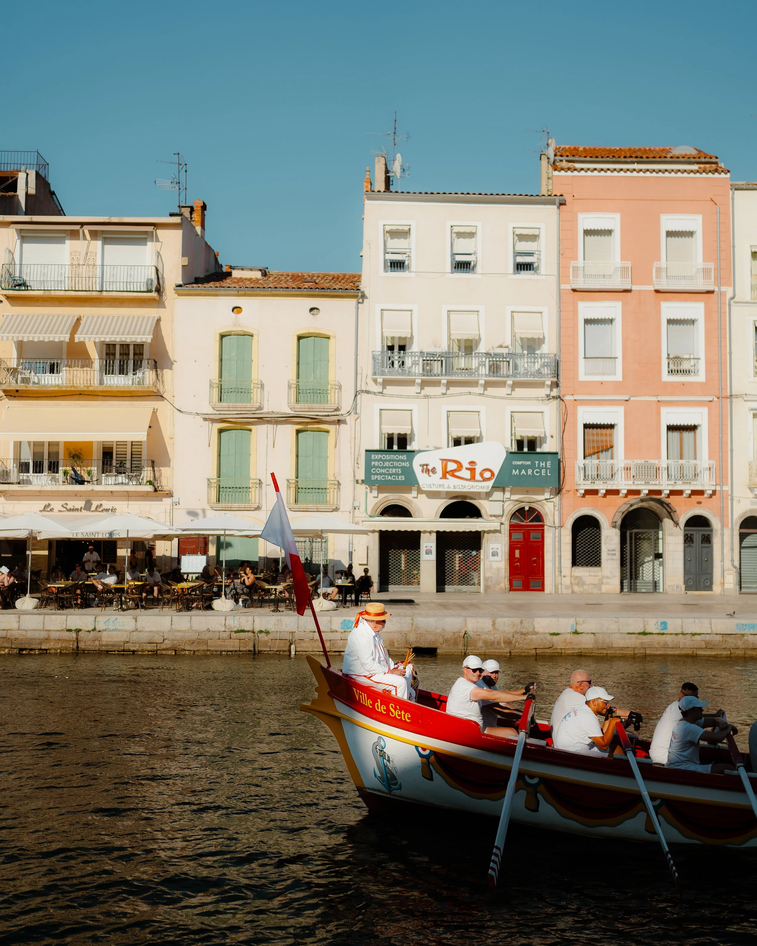 People rowing a boat with a flag on the water in front of colorful buildings with cafes and signs in a European city.