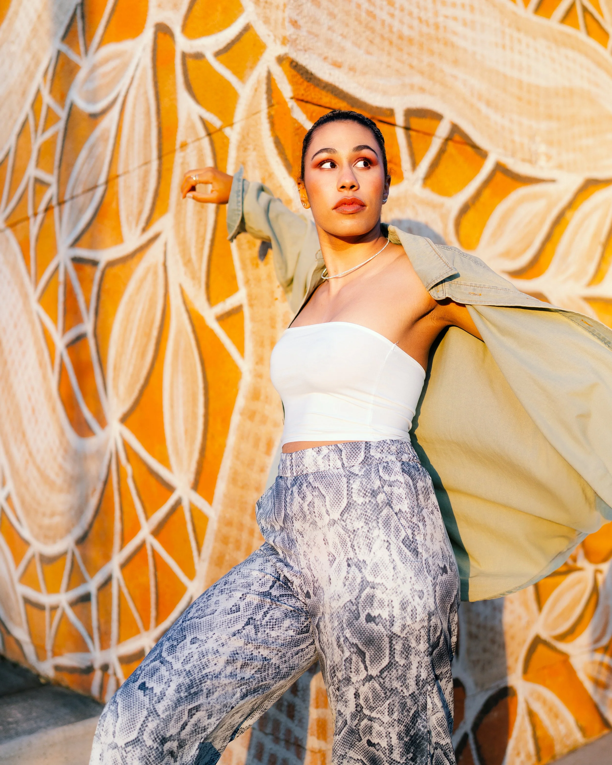 A young woman in a strapless white top, snake-print pants, and an oversized yellow jacket poses in front of a colorful mosaic wall.