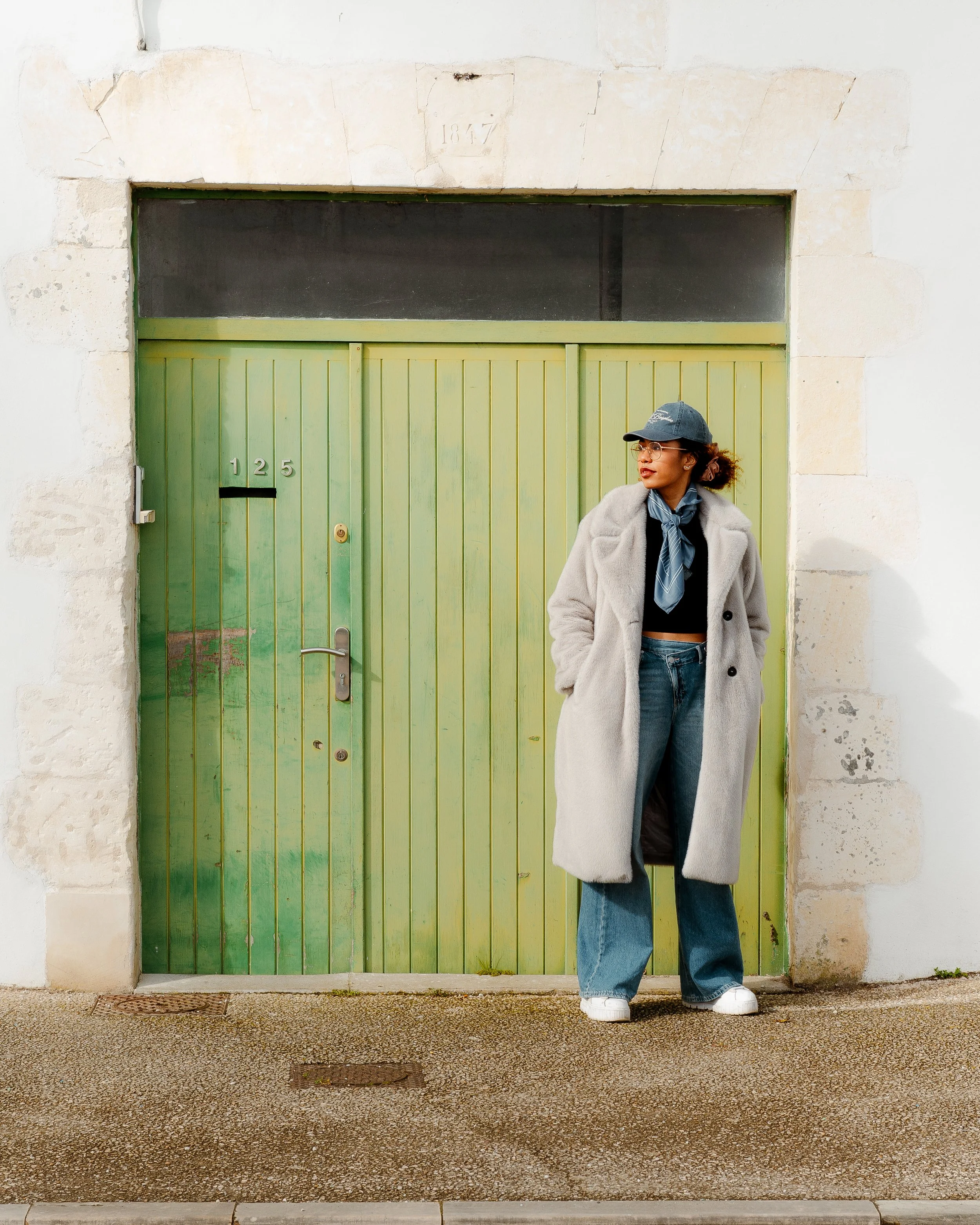 A woman in a grey coat, baseball cap, and jeans standing in front of a green wooden door on a stone building.