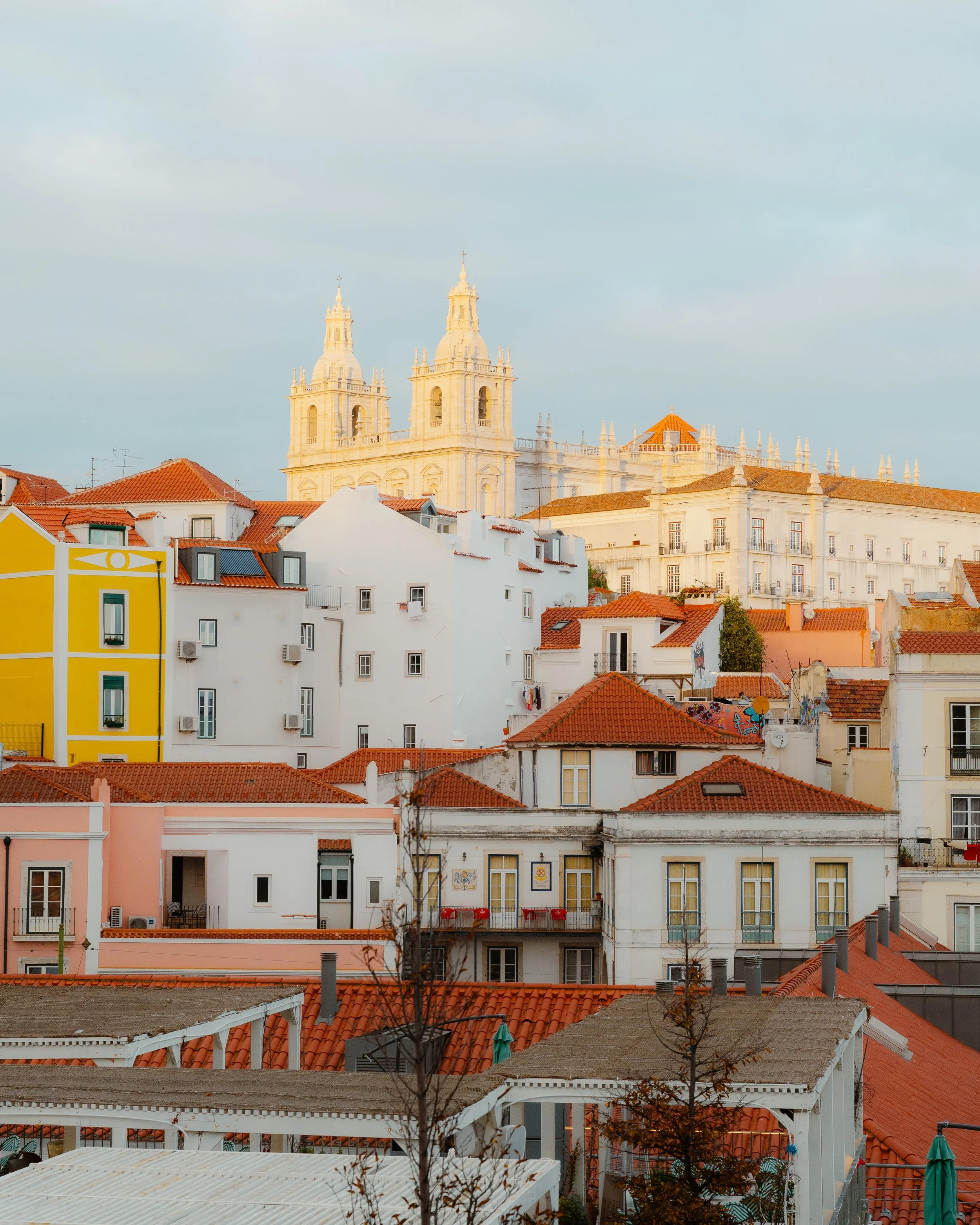 Colorful cityscape of Lisbon, Portugal with white and pastel-colored buildings, red-tiled roofs, and a historic church with twin bell towers in the background.