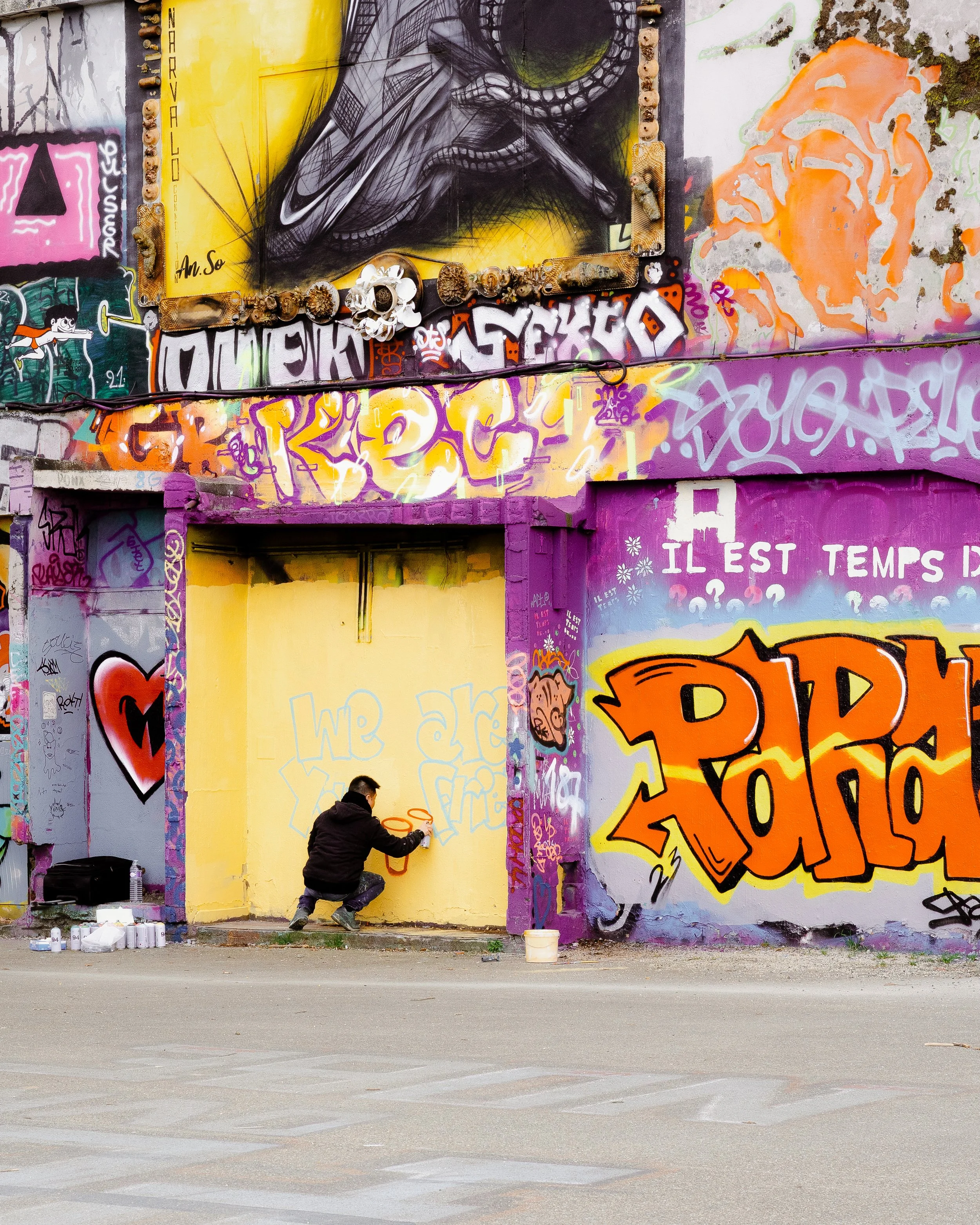 A person spray painting graffiti on a yellow wall in front of a colorful, mural-covered building with various graffiti tags and artwork, including a large orange and yellow piece that says 'POP'.