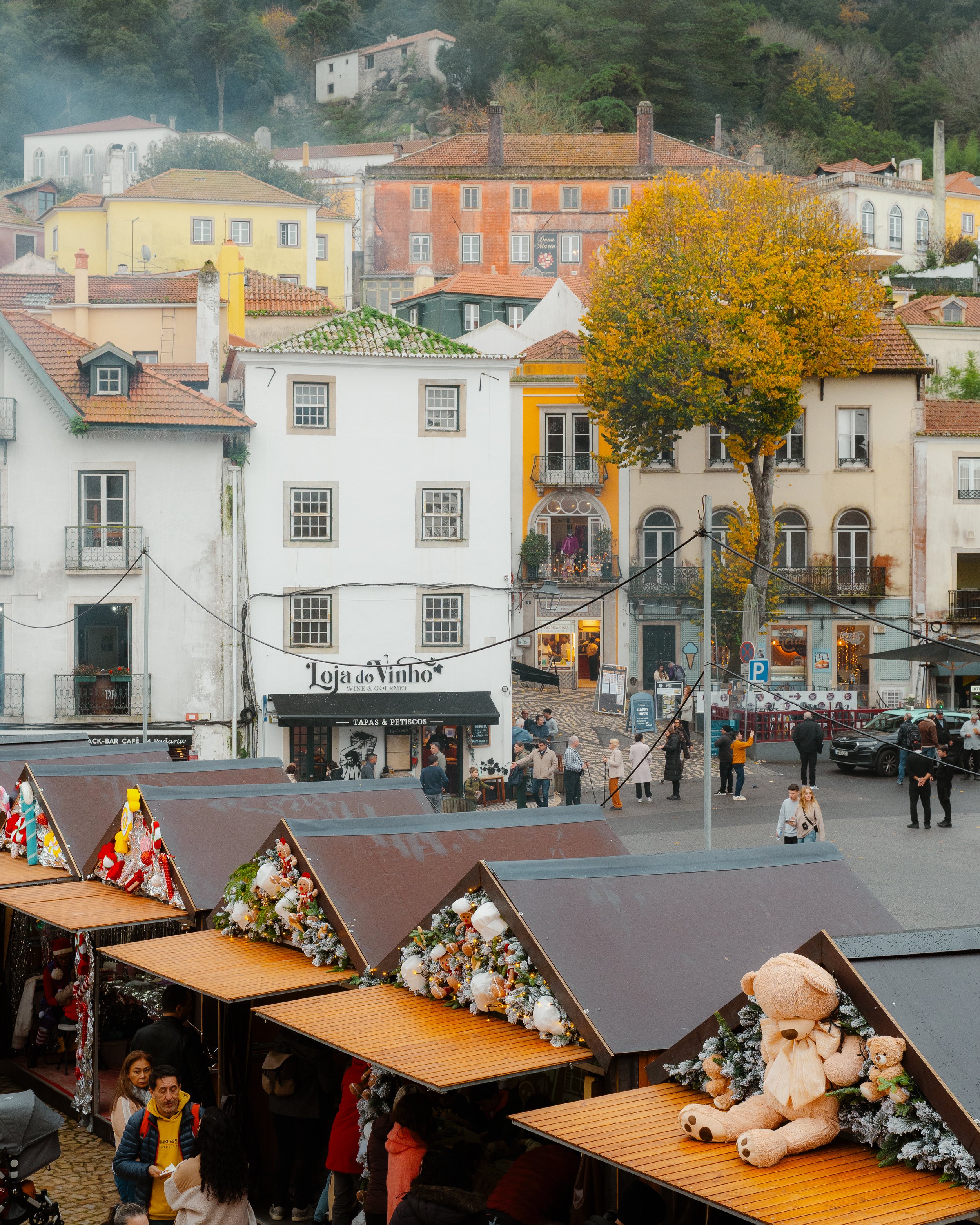 View of a decorated outdoor market with christmas ornaments and stuffed teddy bears, colorful buildings, and a lively crowd in a European village during the holiday season.