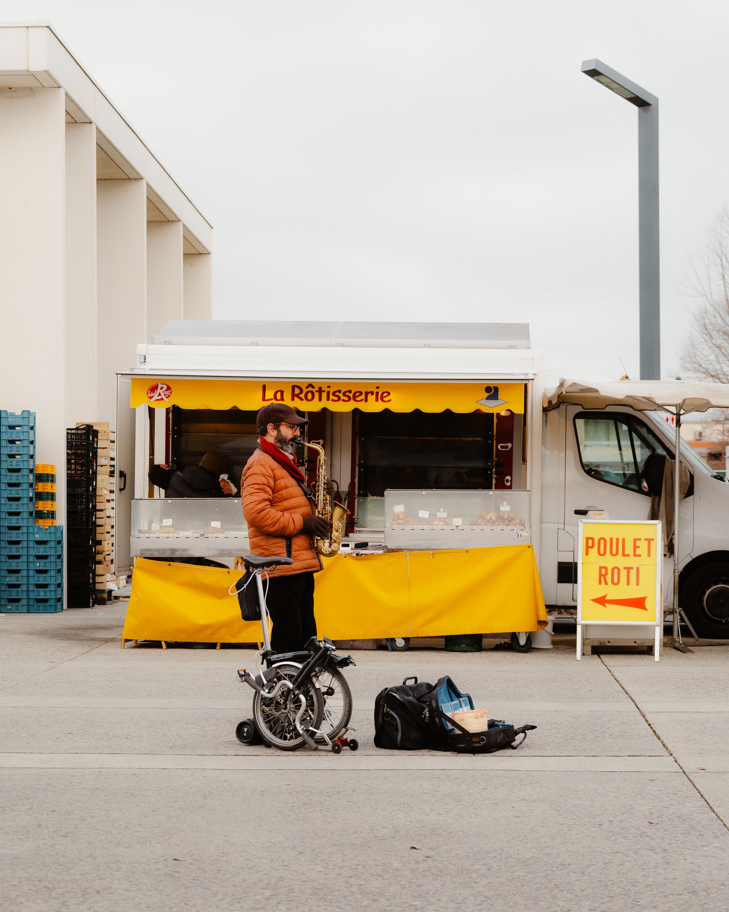 A man with a bass saxophone standing in front of a yellow food truck labeled 'La Rôtisserie' serving roasted chicken and roast beef, with a sign pointing to the food stand.