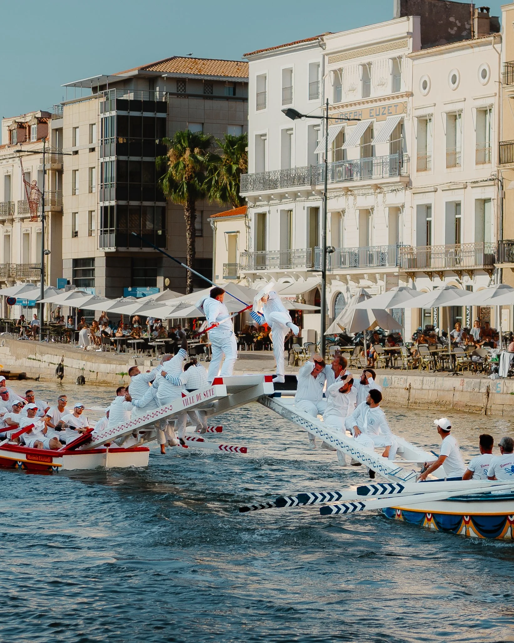 People wearing white clothing participating in a boat race on a river, with some standing on the boat and others sitting, while spectators sit at outdoor tables along the riverbank, and buildings line the background.